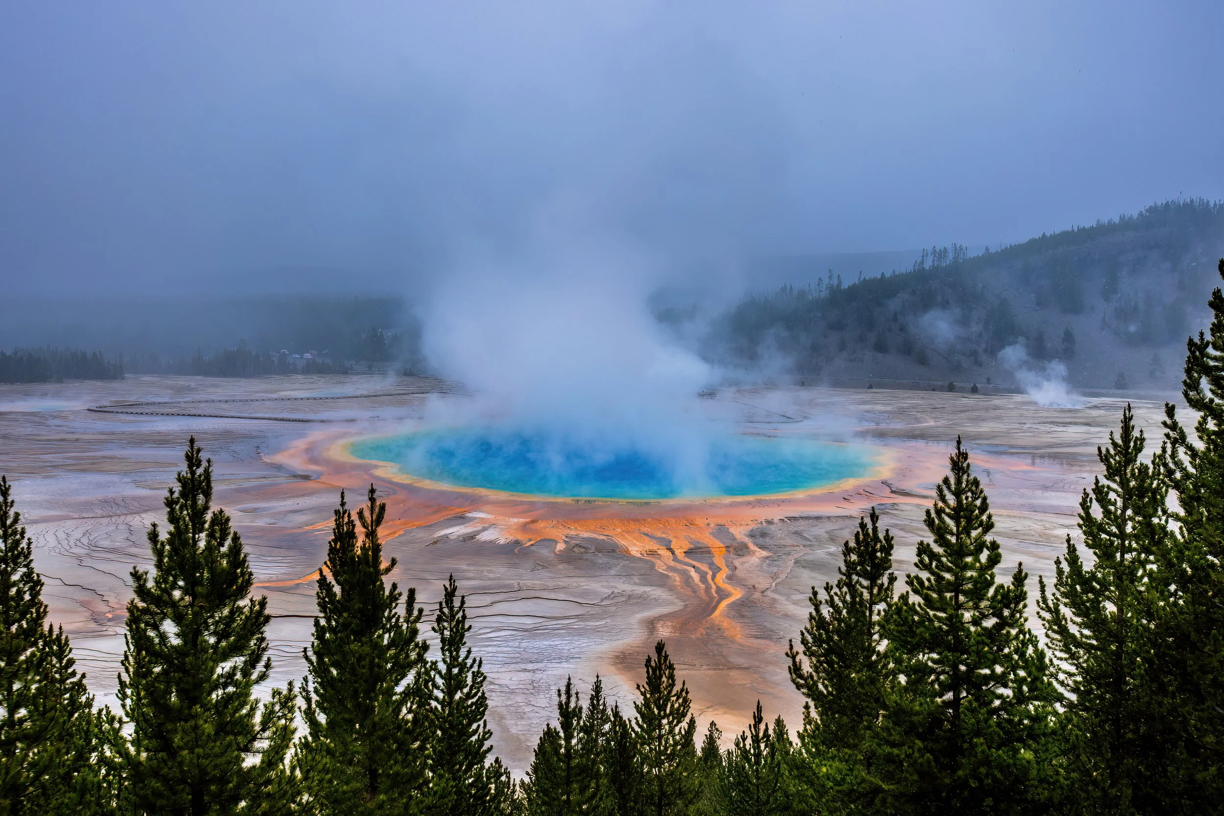 Colorful hot spring pool with steam rising, surrounded by green trees and forested hills under a cloudy sky.