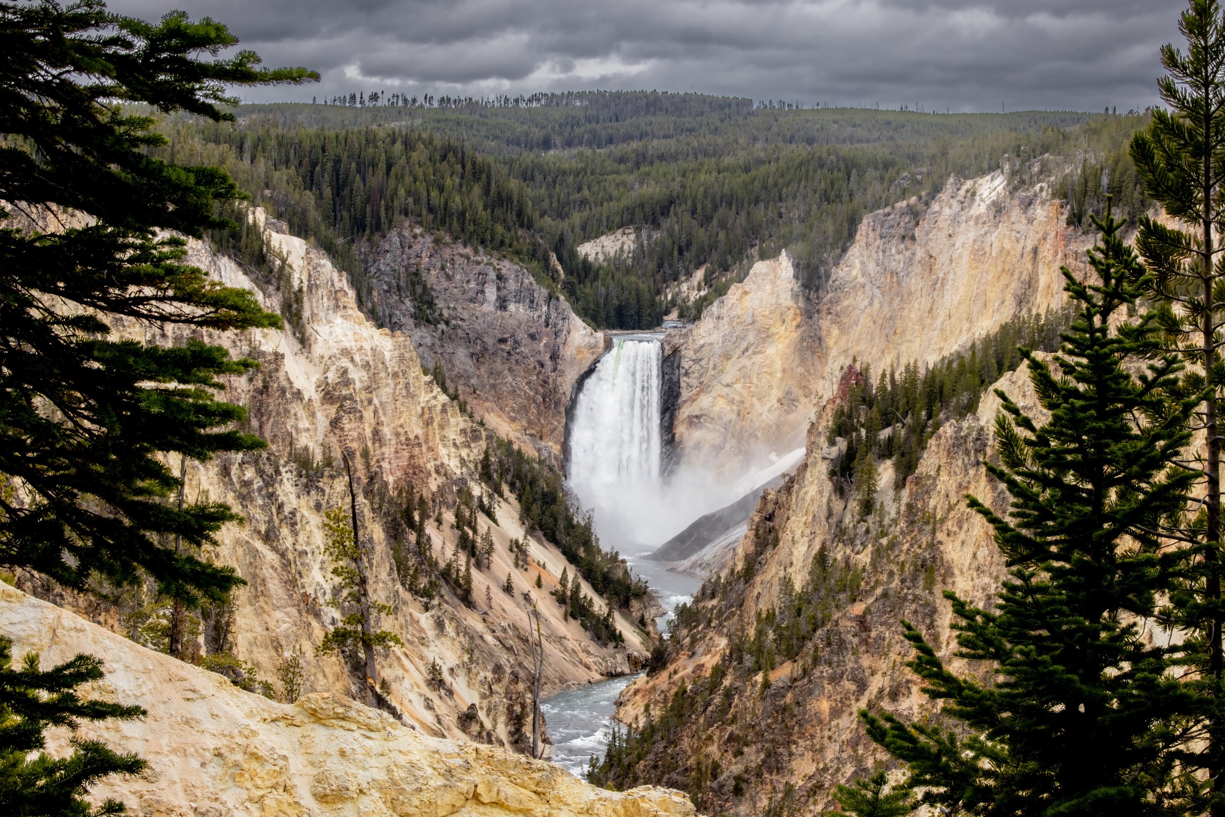 View of a waterfall cascading down a rocky canyon, with a river flowing below and dense evergreen forest on the cliffs, under a cloudy sky.