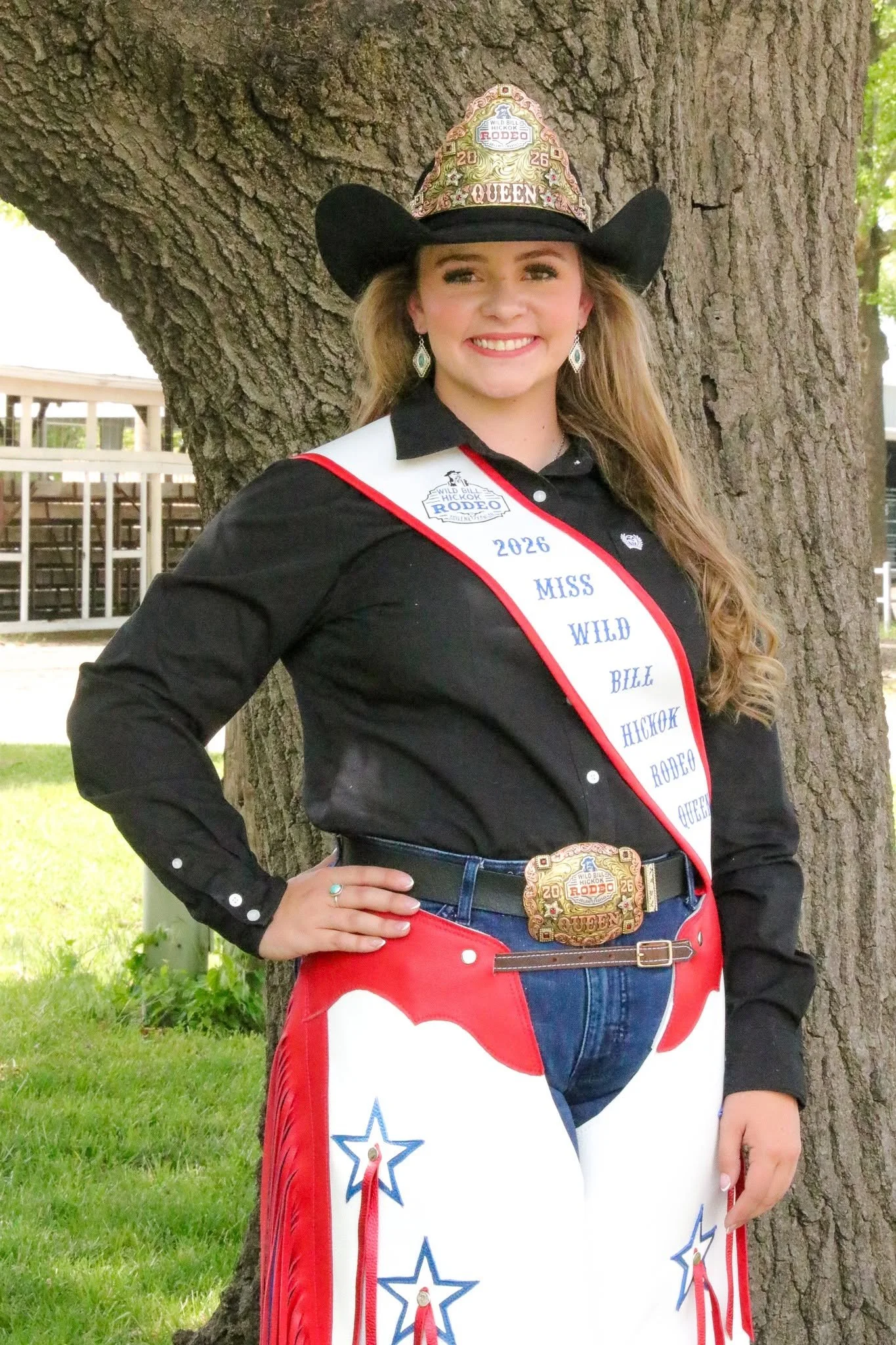 A young woman wearing a cowboy hat with a rodeo logo, a white sash that says '2026 Miss Wild Bill Hickok Rodeo Queen,' and patriotic-colored pants with star designs, standing outdoors against a large tree and a grassy field.