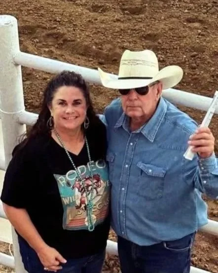 A woman with dark curly hair wearing a black graphic t-shirt and earrings, and an older man with sunglasses, a cowboy hat, and a denim shirt, standing outdoors in front of a white fence.