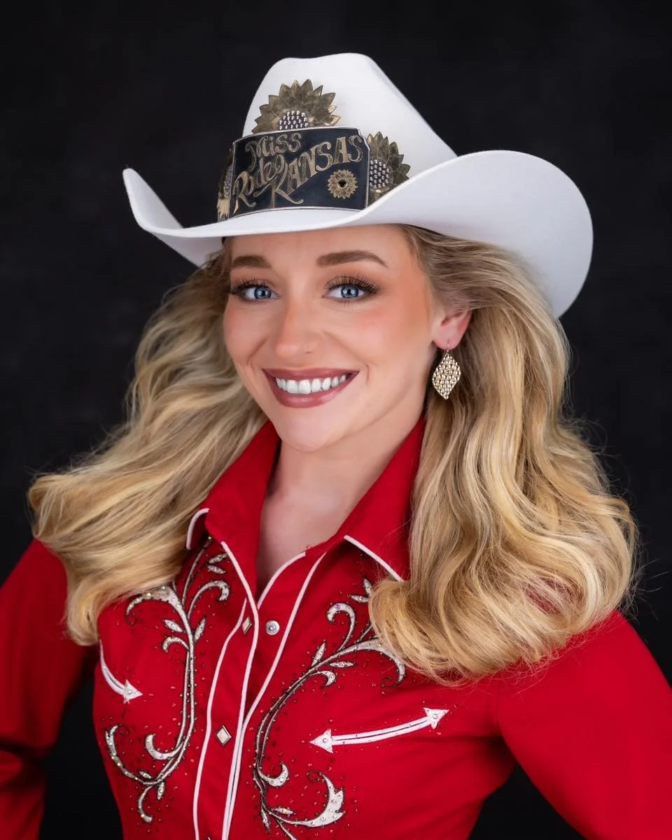 A woman smiling with blonde wavy hair, wearing a white cowboy hat with a decorative badge that reads 'Miss Kansas', a red western shirt with white embroidery, and large gold earrings against a dark background.