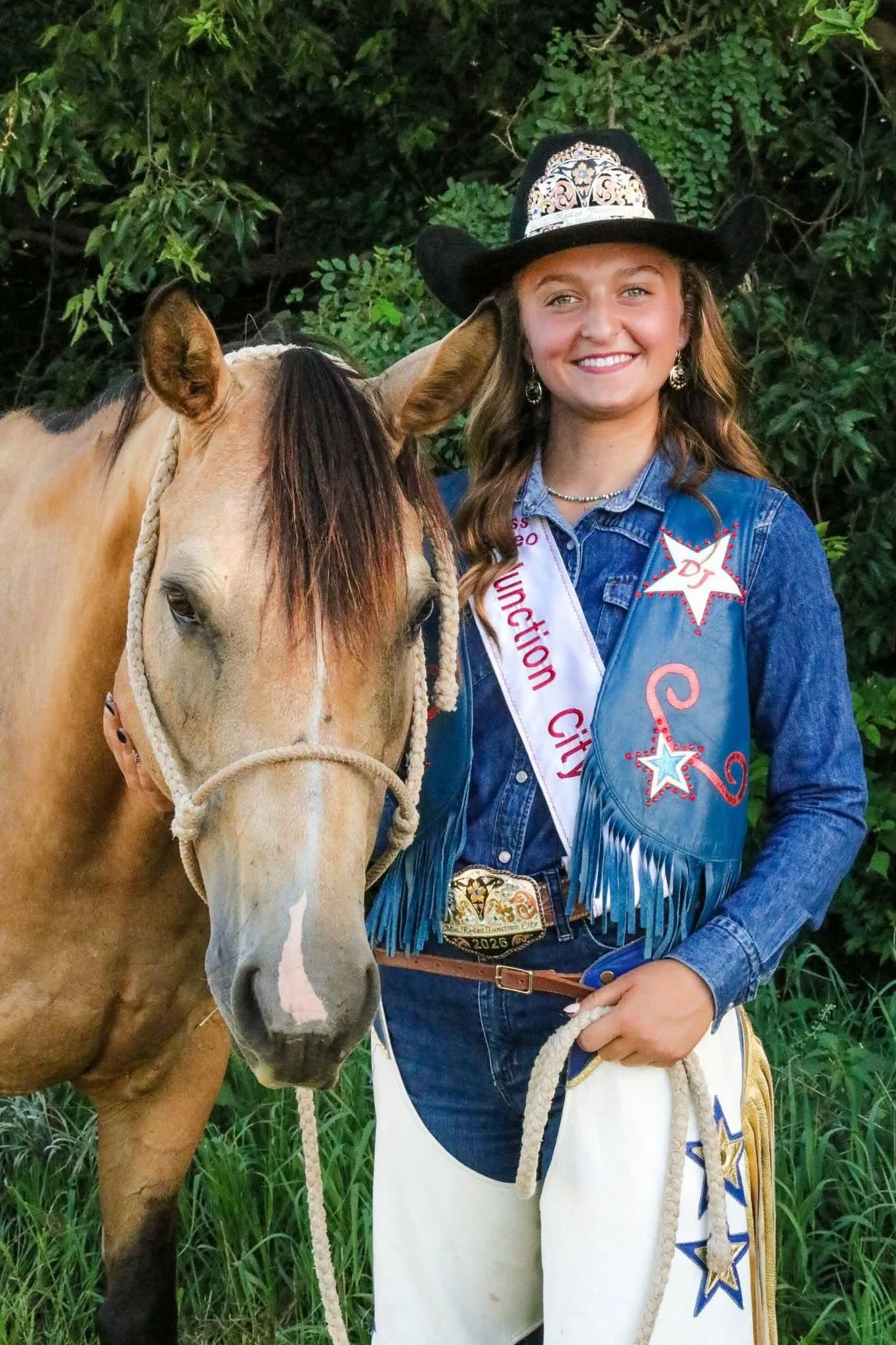 A woman dressed in a cowboy costume, wearing a cowboy hat, denim jacket, and a sash that reads 'Princess,' standing next to a light-colored horse with a braided mane, outdoors with green foliage in the background.