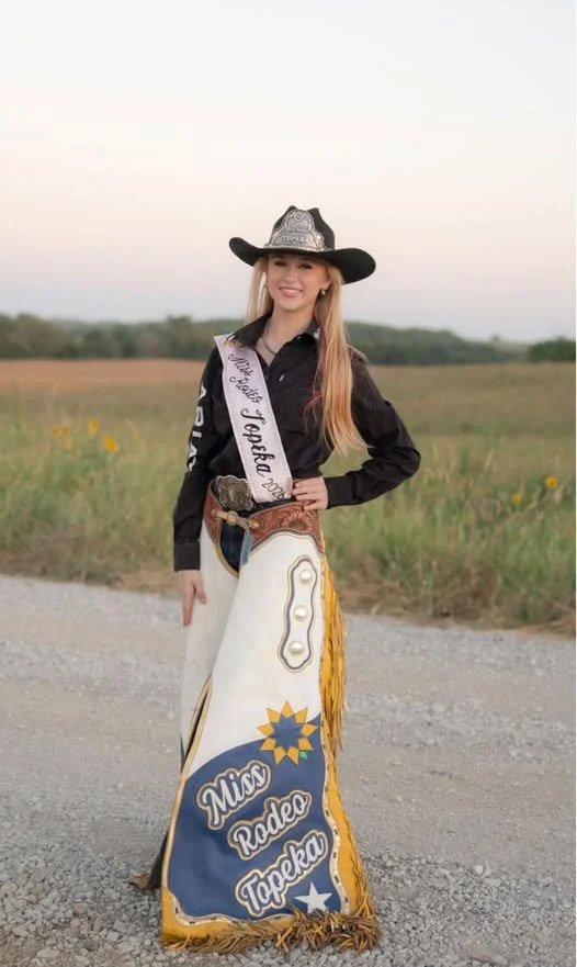 Young woman in cowboy hat and western attire standing outdoors in a rural area, wearing a sash that reads 'Miss Rodeo Topeka' and a decorative rodeo skirt.
