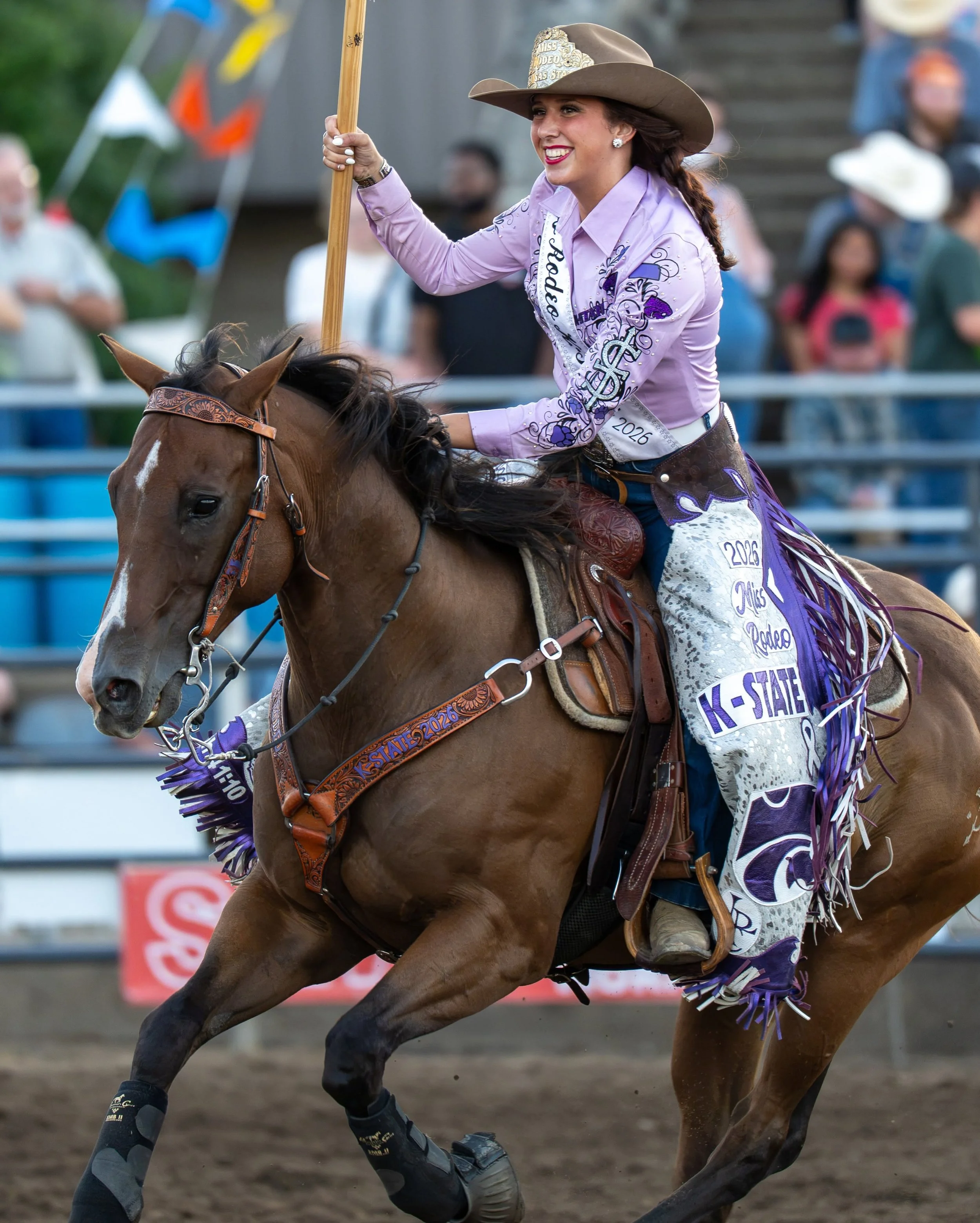 A woman riding a horse during a rodeo event, wearing a cowboy hat and a purple shirt, holding a flag with a smile on her face.