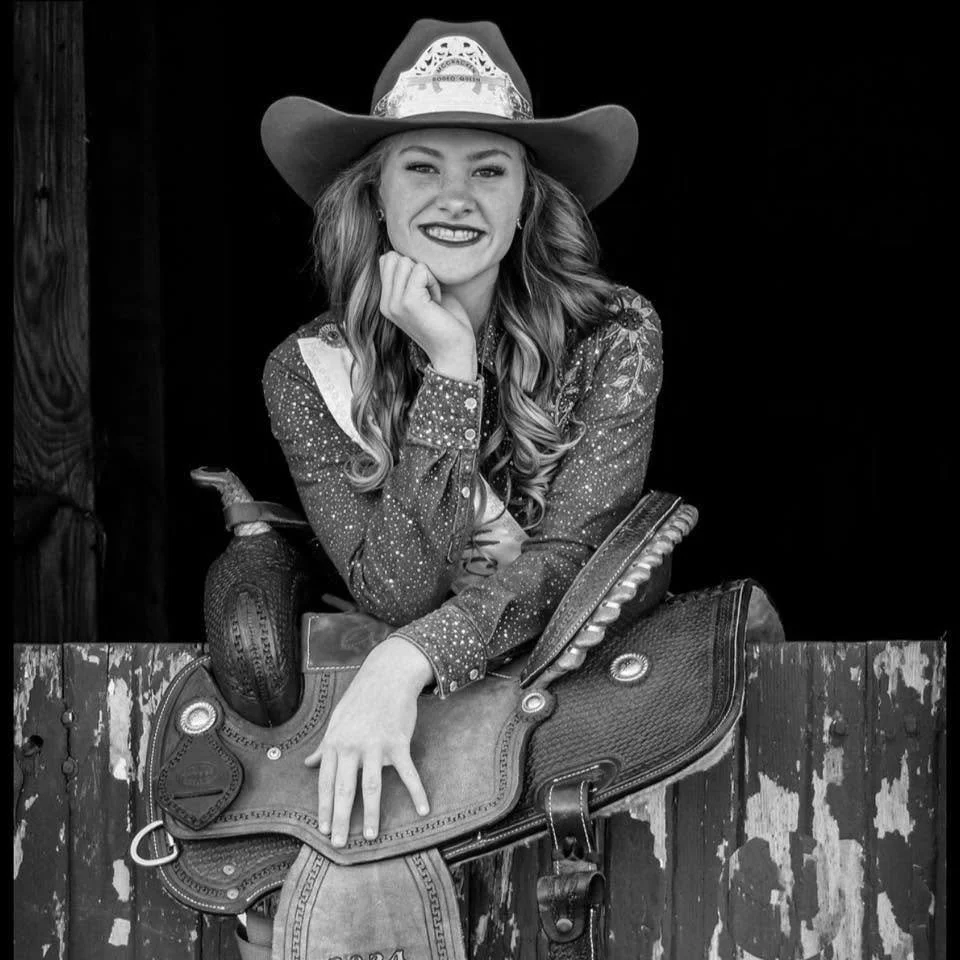 A woman smiling, wearing a cowboy hat and a patterned shirt, sitting behind a wooden fence, with a saddle in front of her.
