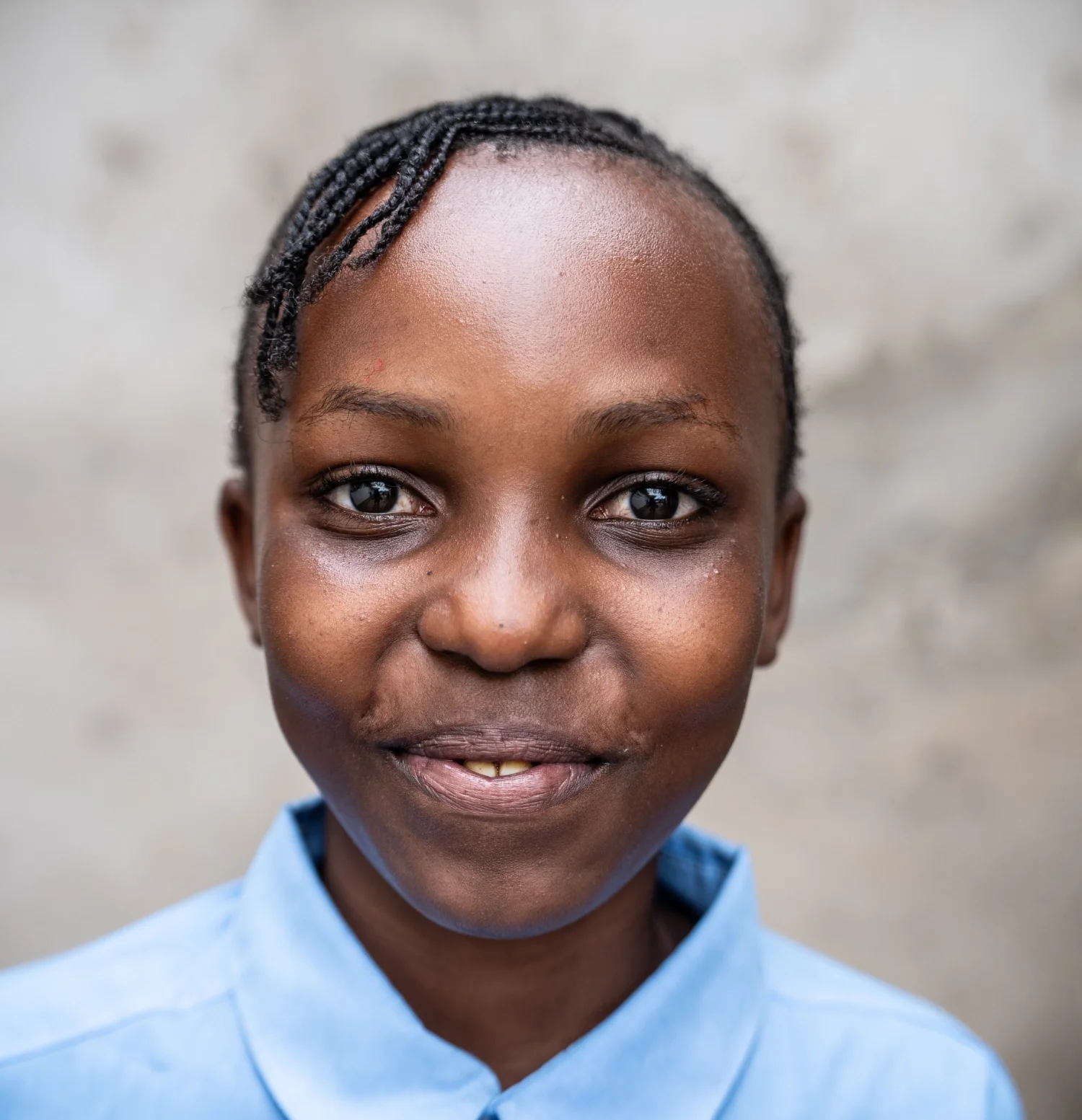 Close-up of a young girl with braided hair, smiling, wearing a light blue collared shirt, standing against a neutral background.