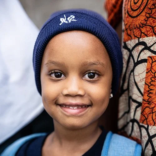 A young girl with a blue knit cap smiling at the camera, standing next to someone wearing a patterned orange and black shirt.
