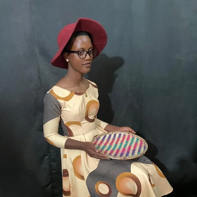A young girl wearing a red wide-brimmed hat, glasses, and earrings, dressed in a long patterned dress, seated against a dark background, holding a colorful woven tray.