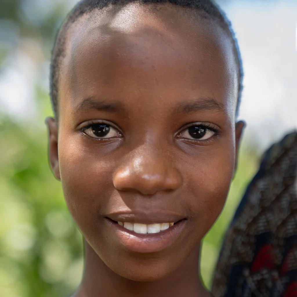 Close-up of a smiling young girl with short hair and dark skin, outdoors with blurred greenery in the background.
