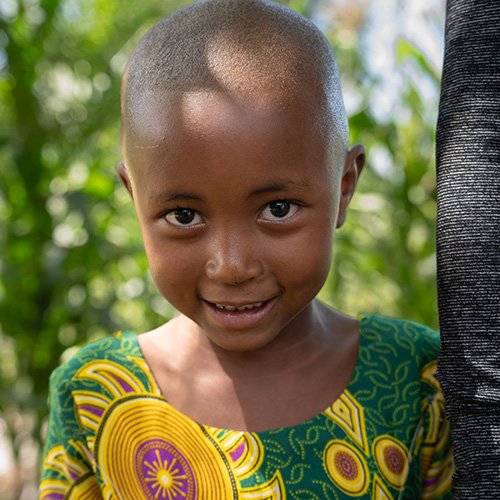 A young girl with a shaved head smiling outdoors with greenery in the background.