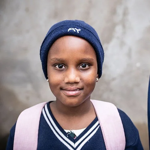 A young girl with dark skin wearing a navy blue beanie hat, a navy blue sweater with white stripes, and a light pink backpack, standing outdoors in front of a gray wall.