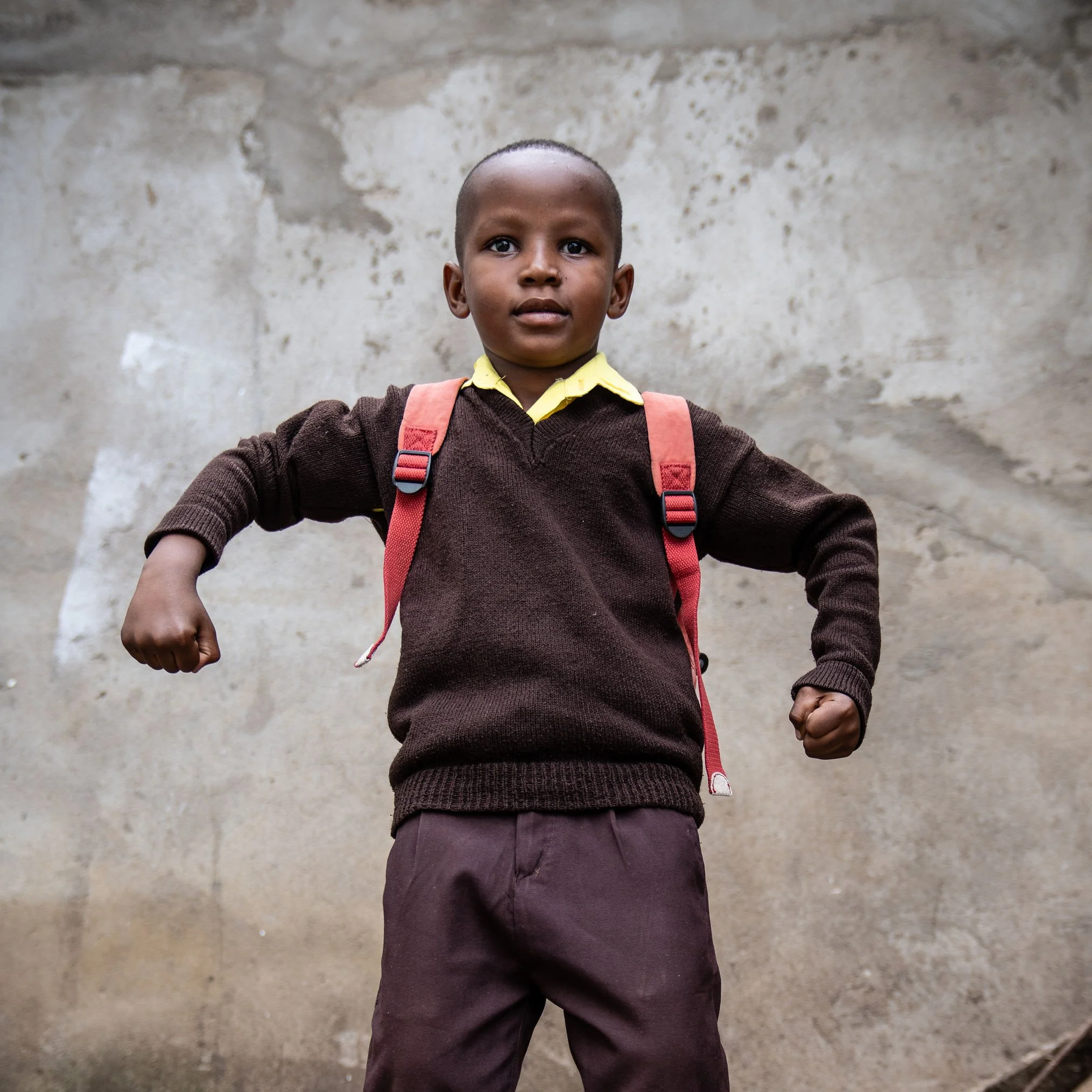 A young boy with a shaved head, wearing a yellow shirt, brown sweater, and maroon pants, standing outdoors in front of a weathered concrete wall, with a red backpack.