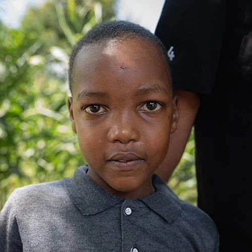 Close-up of a young boy with short hair and a serious expression, standing outdoors with greenery in the background.