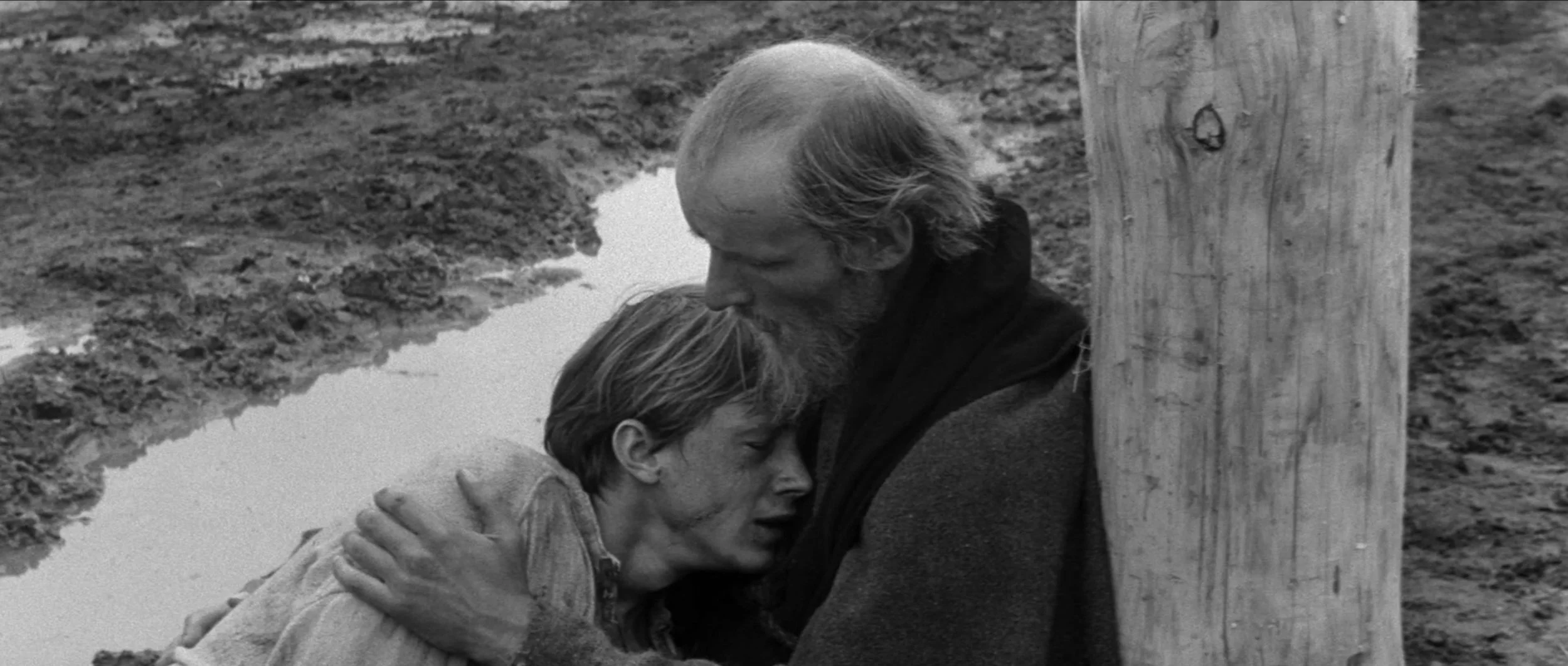 A black-and-white photo of a man and a boy embracing behind a wooden post, with muddy ground and a puddle in the background.