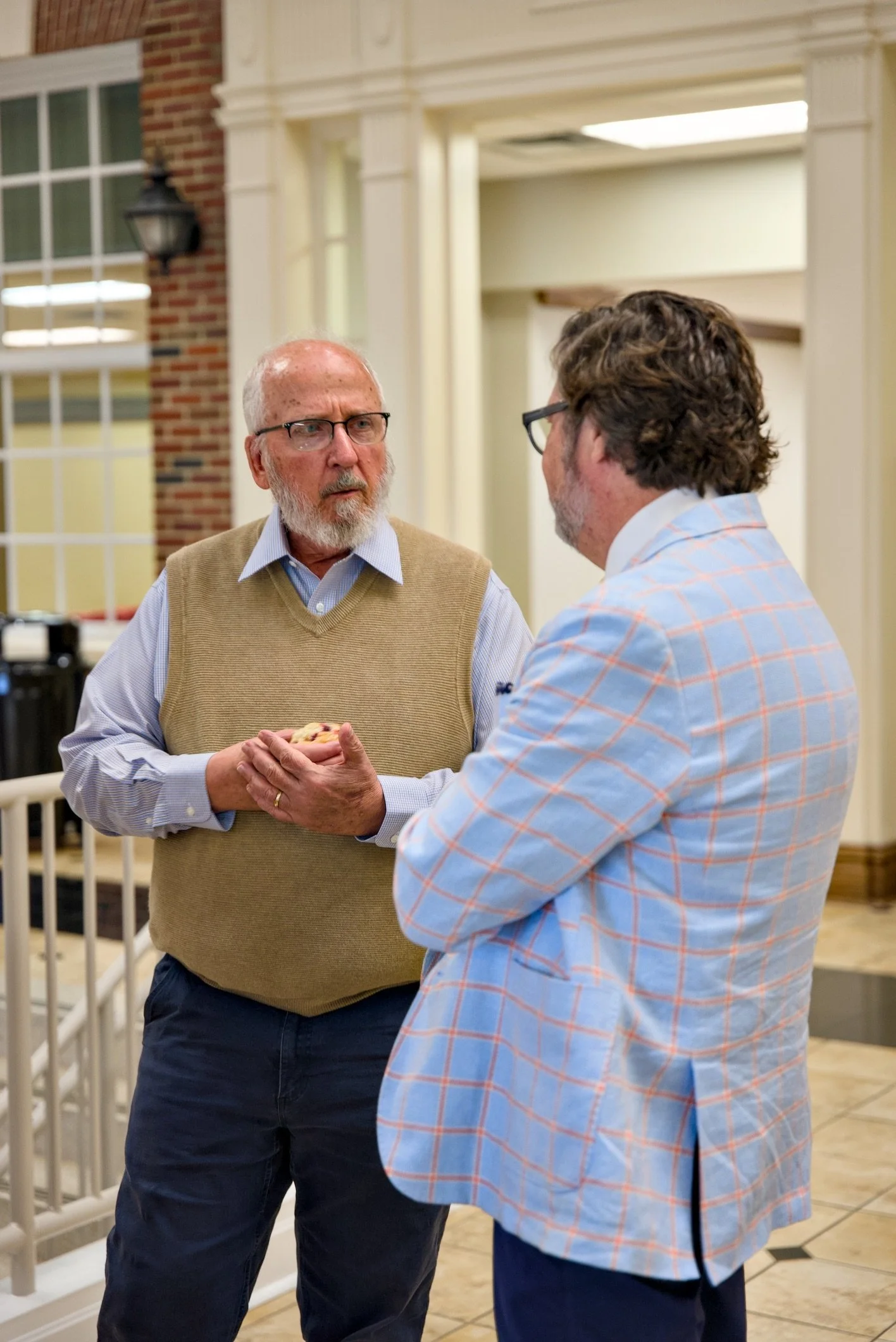 Two men are engaged in conversation indoors. One man, with a beard and glasses, is wearing a beige vest over a light blue shirt. The other man, with curly hair and glasses, is wearing a plaid blazer. They are standing near a staircase.