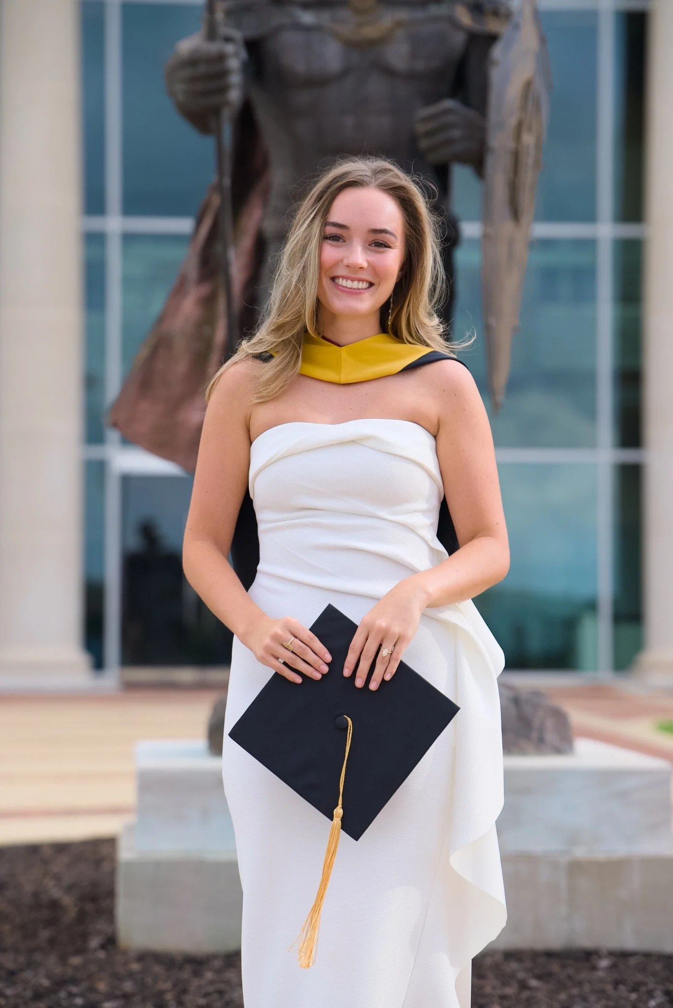 A young woman in a white strapless dress holding a graduation cap with a gold tassel, standing outdoors in front of a building with large glass windows and a large statue of a knight in armor in the background. She is smiling.