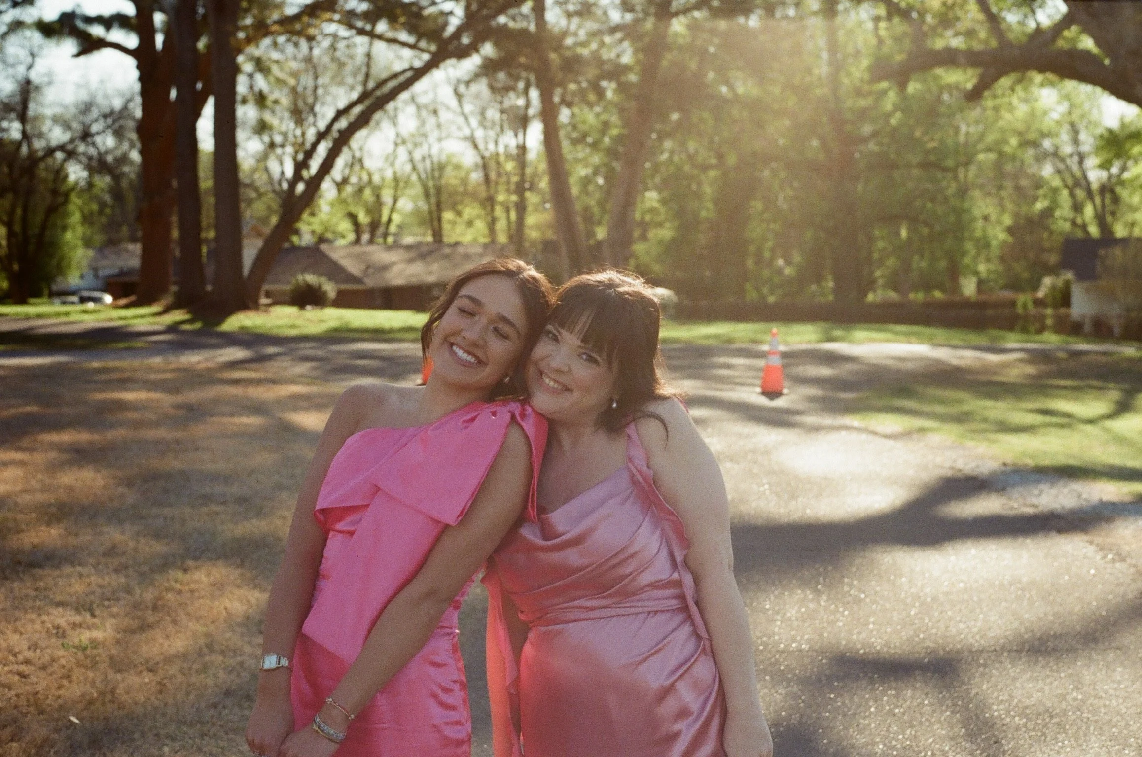 Two women wearing pink dresses embracing and smiling outdoors. Background features trees and a house with a driveway, sunny weather lighting.