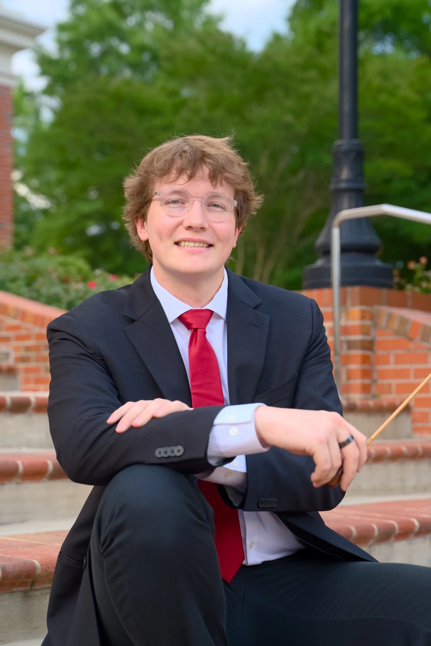 A young man with light brown curly hair, wearing a dark suit, white shirt, and red tie, sitting outdoors on brick stairs, smiling with his arms crossed, with greenery and a black lamp post in the background.
