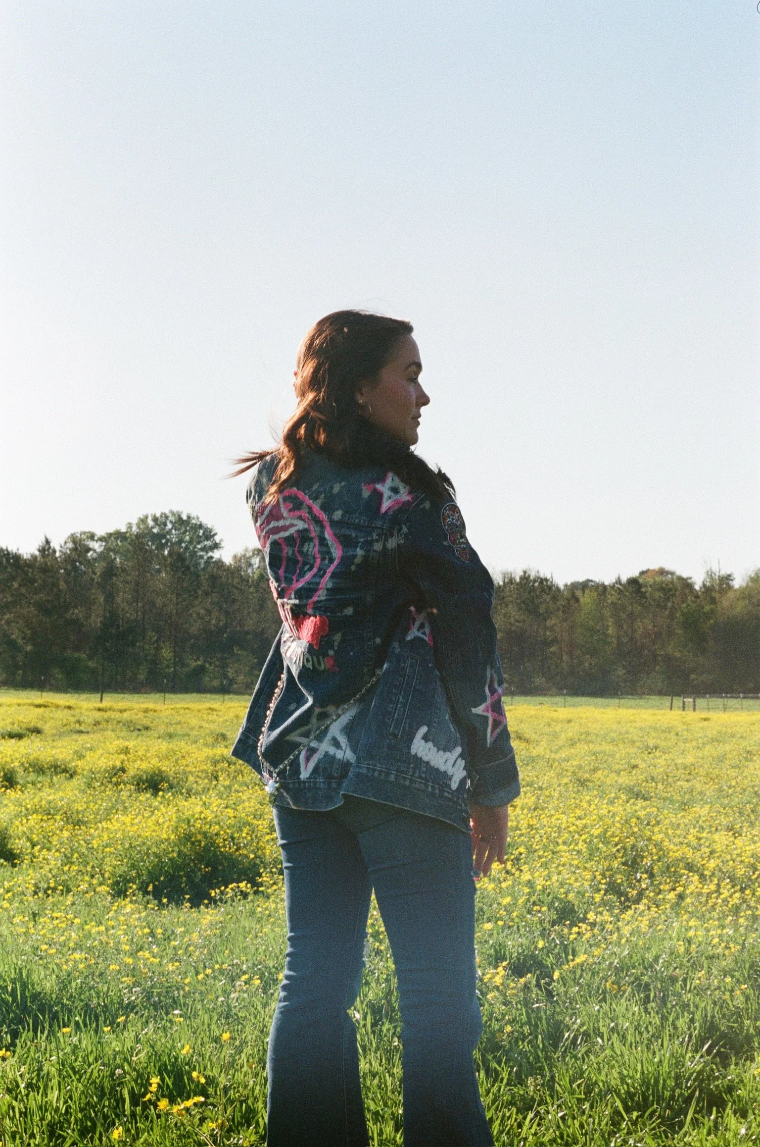 A woman with dark hair in a ponytail standing in a field of yellow flowers, wearing a black jacket with colorful patches and blue jeans, with trees in the background and a clear sky.