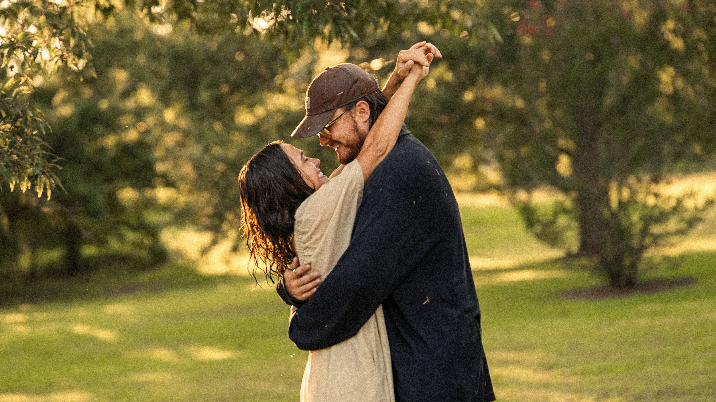 A couple is dancing outdoors during sunset, with the man lifting the woman in his arms. They are smiling and looking into each other's eyes, surrounded by trees and a grassy area.