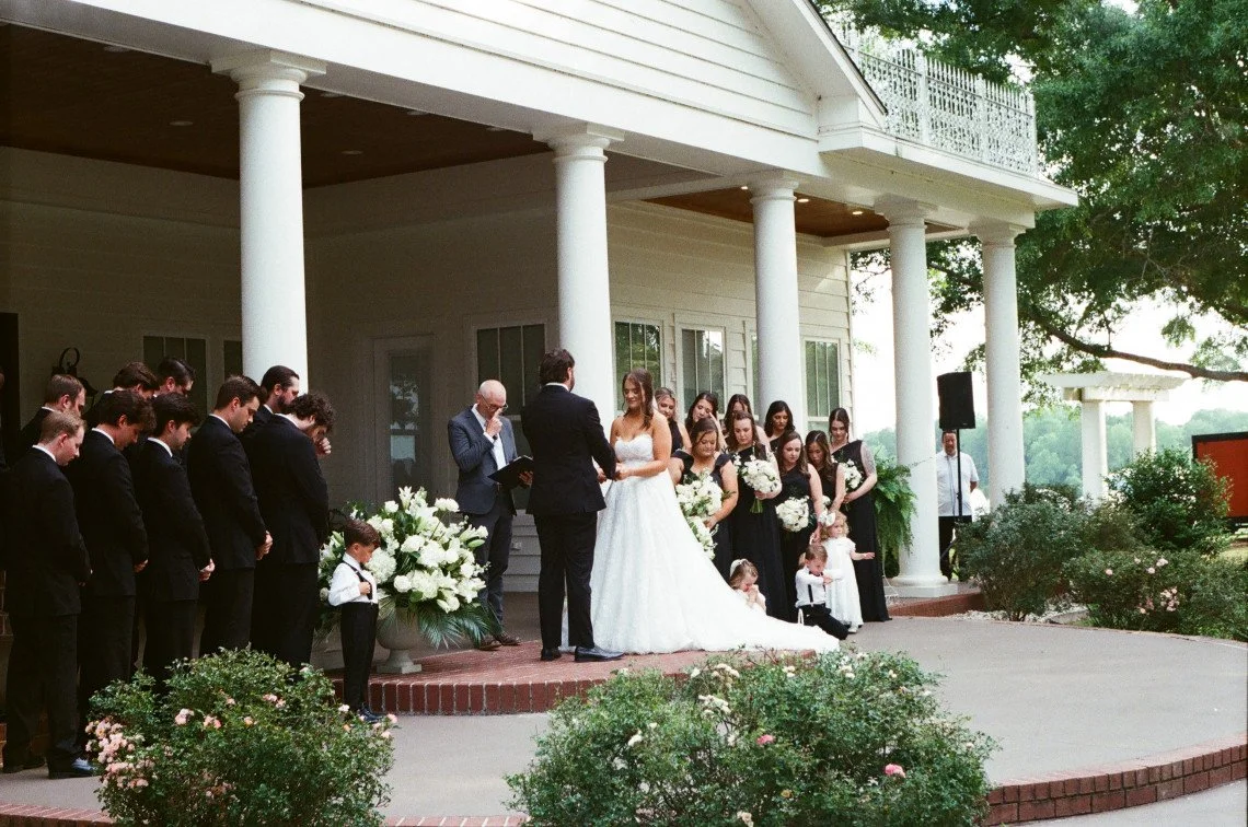 Wedding ceremony taking place on a porch with white pillars, a bride and groom standing in front of an officiant, surrounded by bridesmaids and groomsmen, with children in formal attire, outdoor setting with greenery.