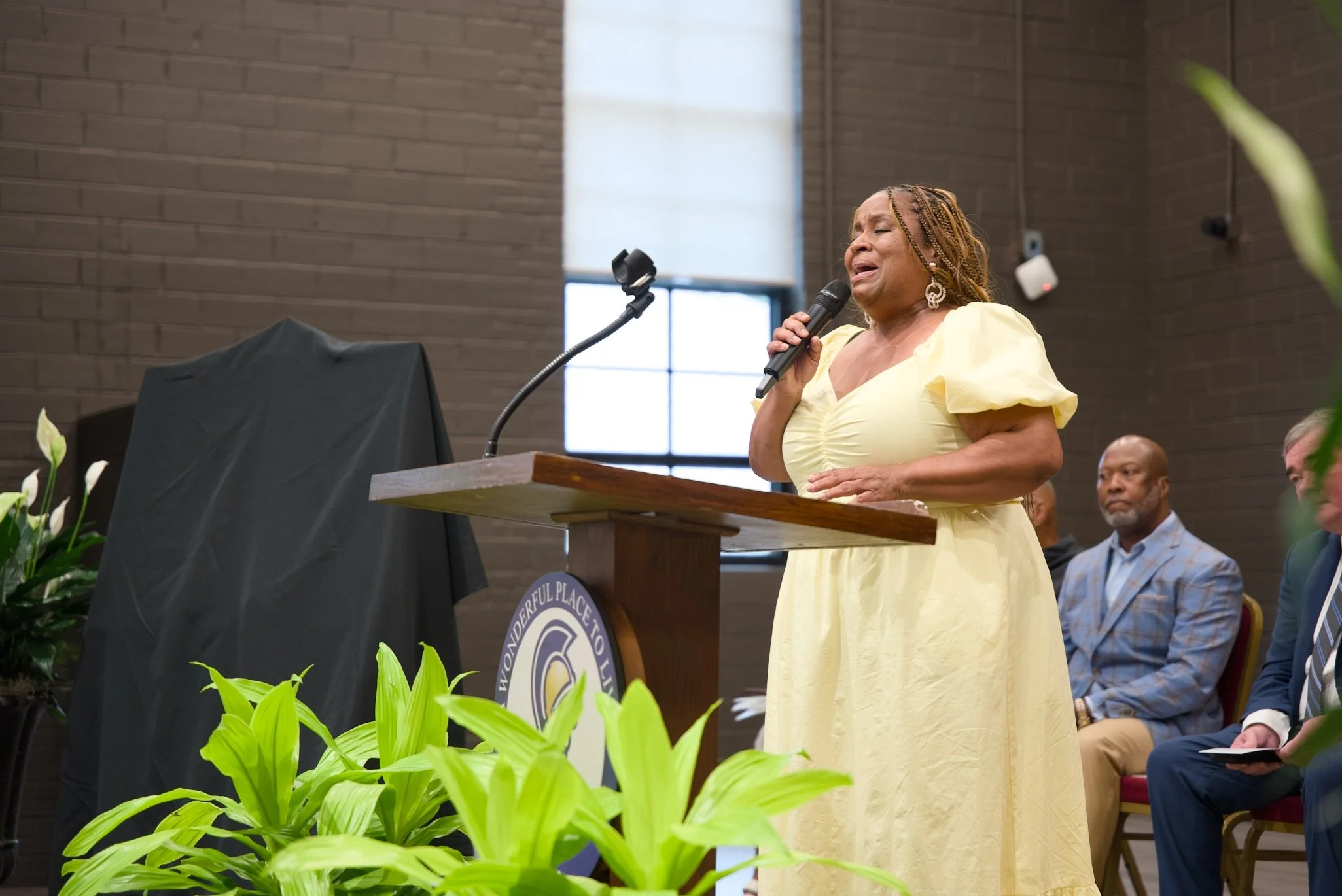 A woman in a yellow dress singing into a microphone at a podium during an indoor event, with seated attendees in the background and green plants in the foreground.