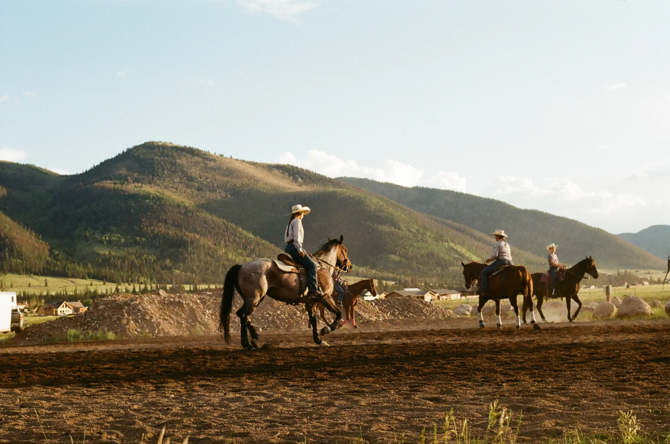 A group of people riding horses in a field with mountains in the background and a few houses nearby, during the daytime.