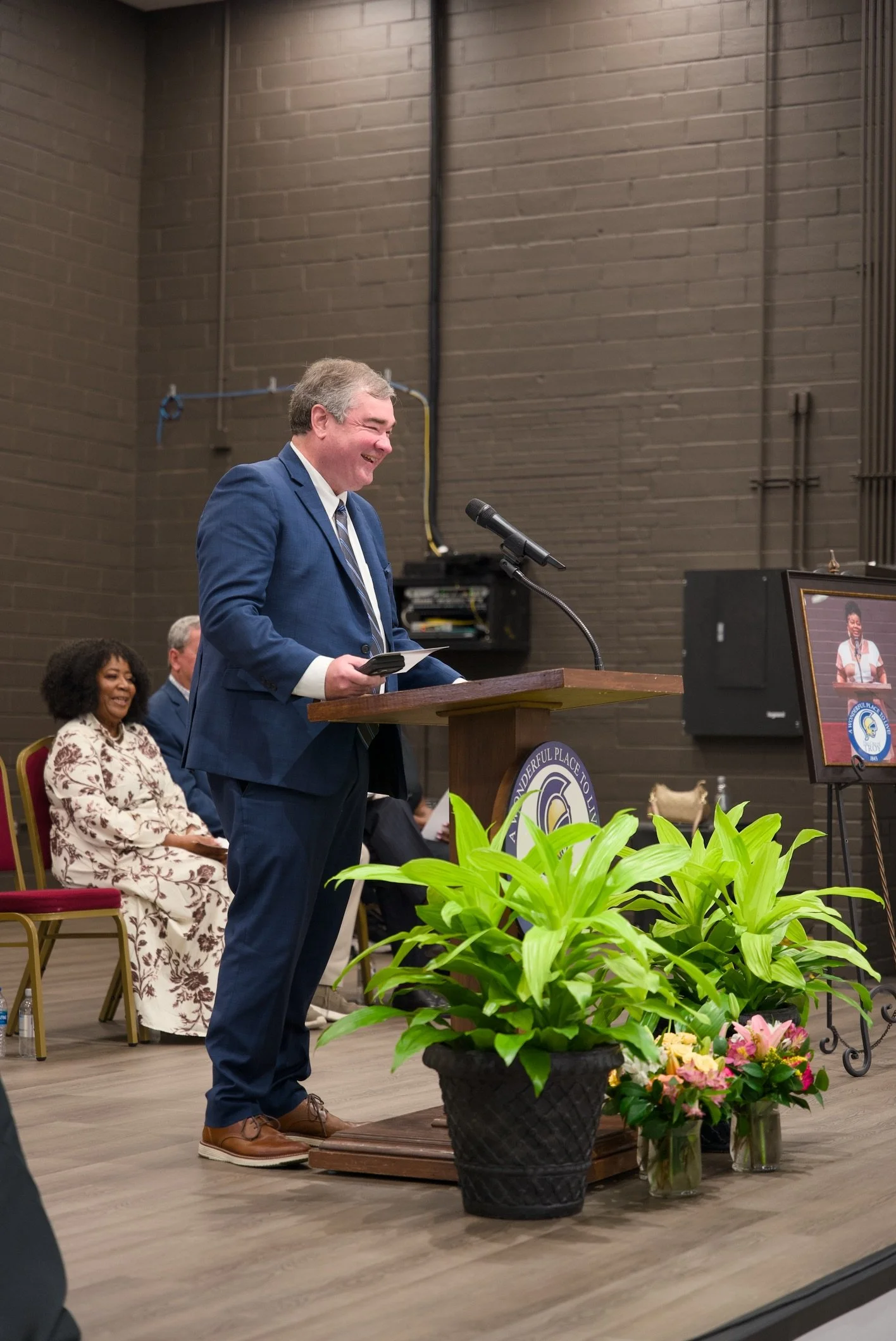 A man in a blue suit is standing at a podium, speaking into a microphone during an indoor event. Behind him, a woman in a floral dress is sitting and smiling. There are bright green plants and flowers in front of the podium, and a framed photo is dis