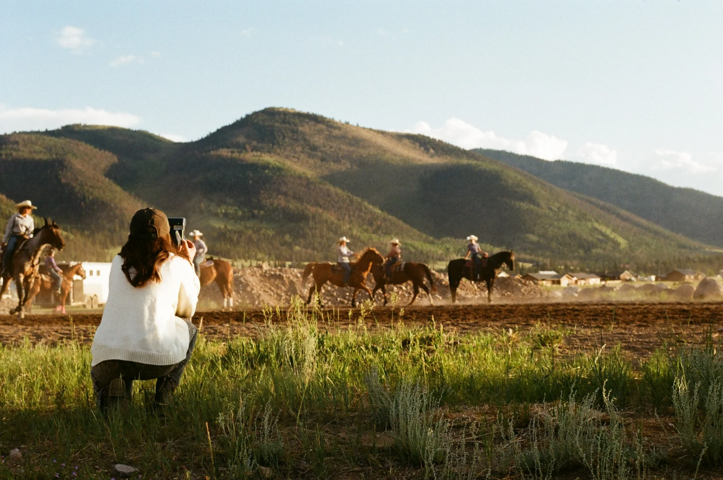 A woman kneeling on grass taking a photo of a group of people riding horses in a field with mountains in the background.