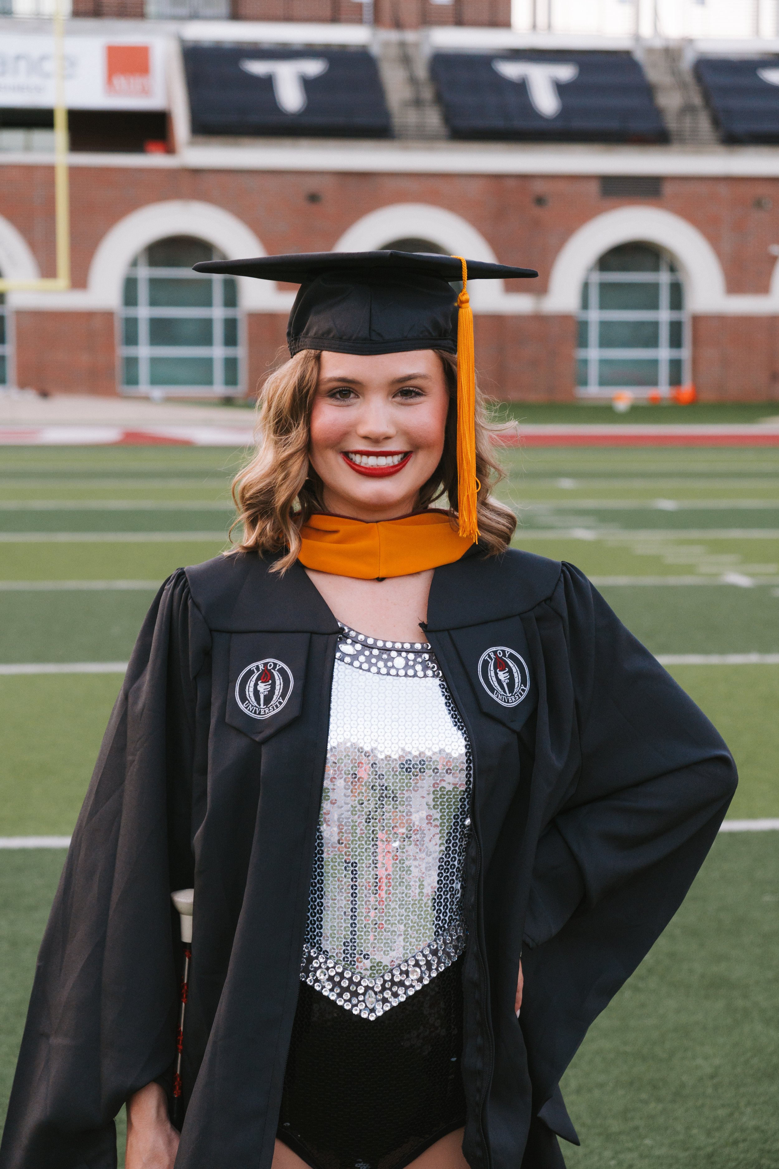 Young woman in graduation attire, including a black cap with a yellow tassel and a gown, standing on a football field.