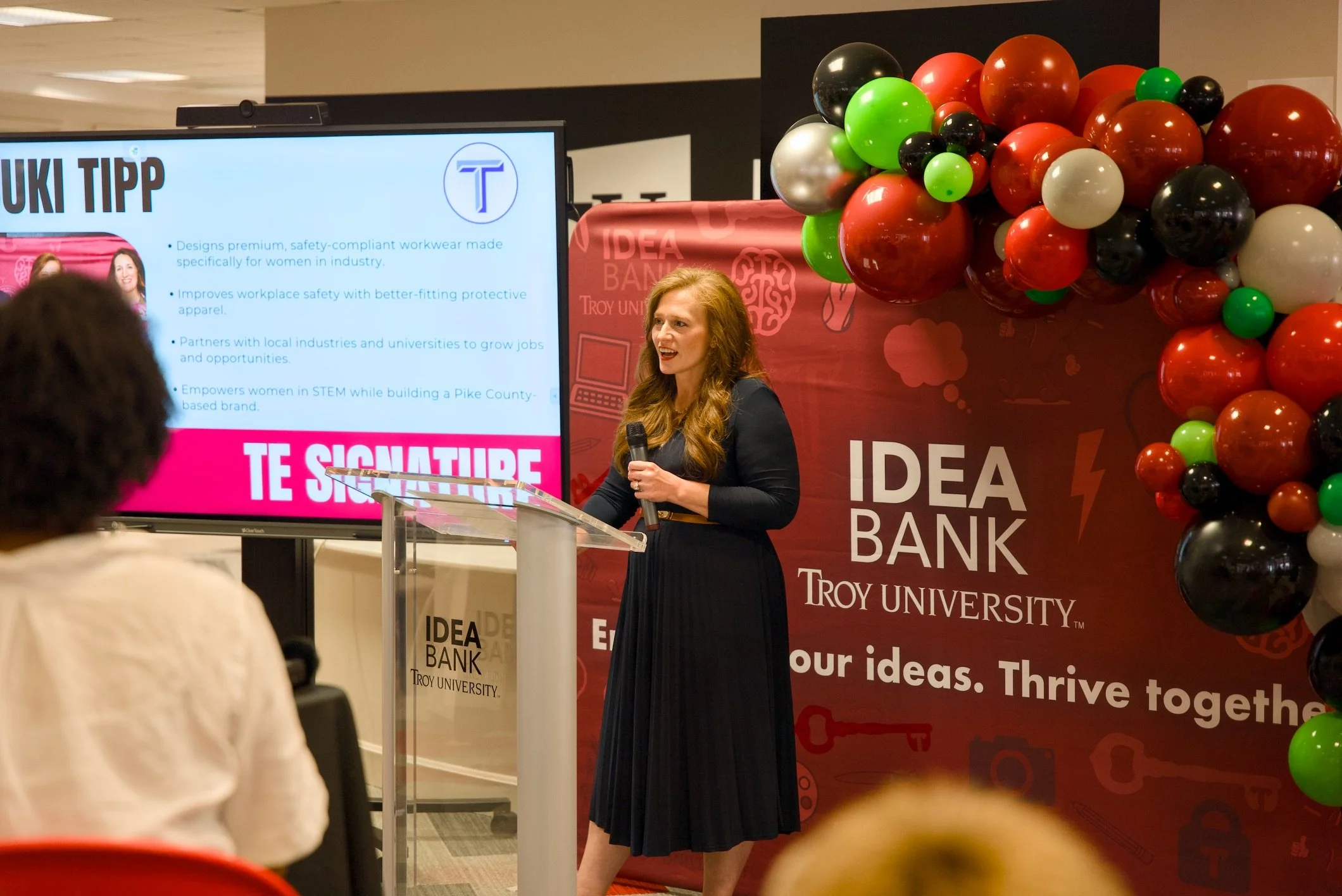 A woman with long red hair is speaking at a podium during a presentation at Troy University. Behind her is a large cluster of colorful balloons and a screening screen displaying a slide titled 'TUKI TIPP.'