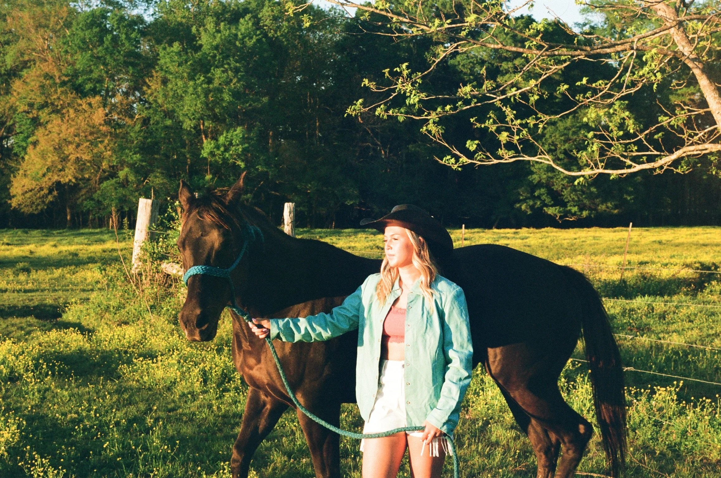 A young woman with long blonde hair, wearing a black cowboy hat, a light green jacket, and shorts, stands next to a brown horse in a grassy field surrounded by trees during the daytime.