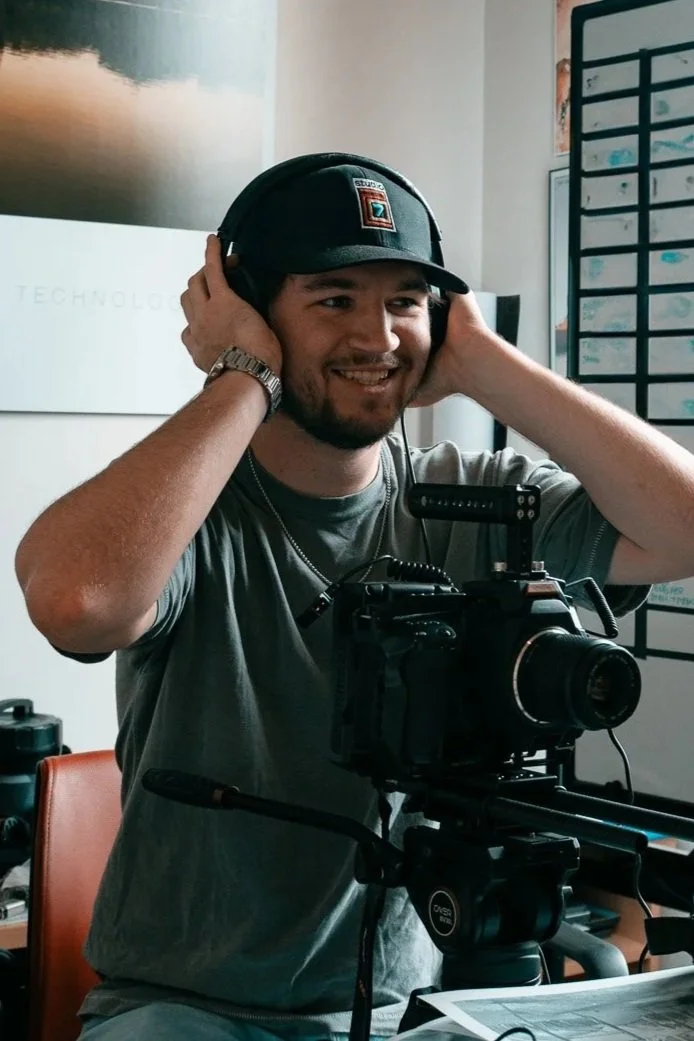 Man adjusting headphones, smiling, with a camera on a tripod, in an indoor office environment.