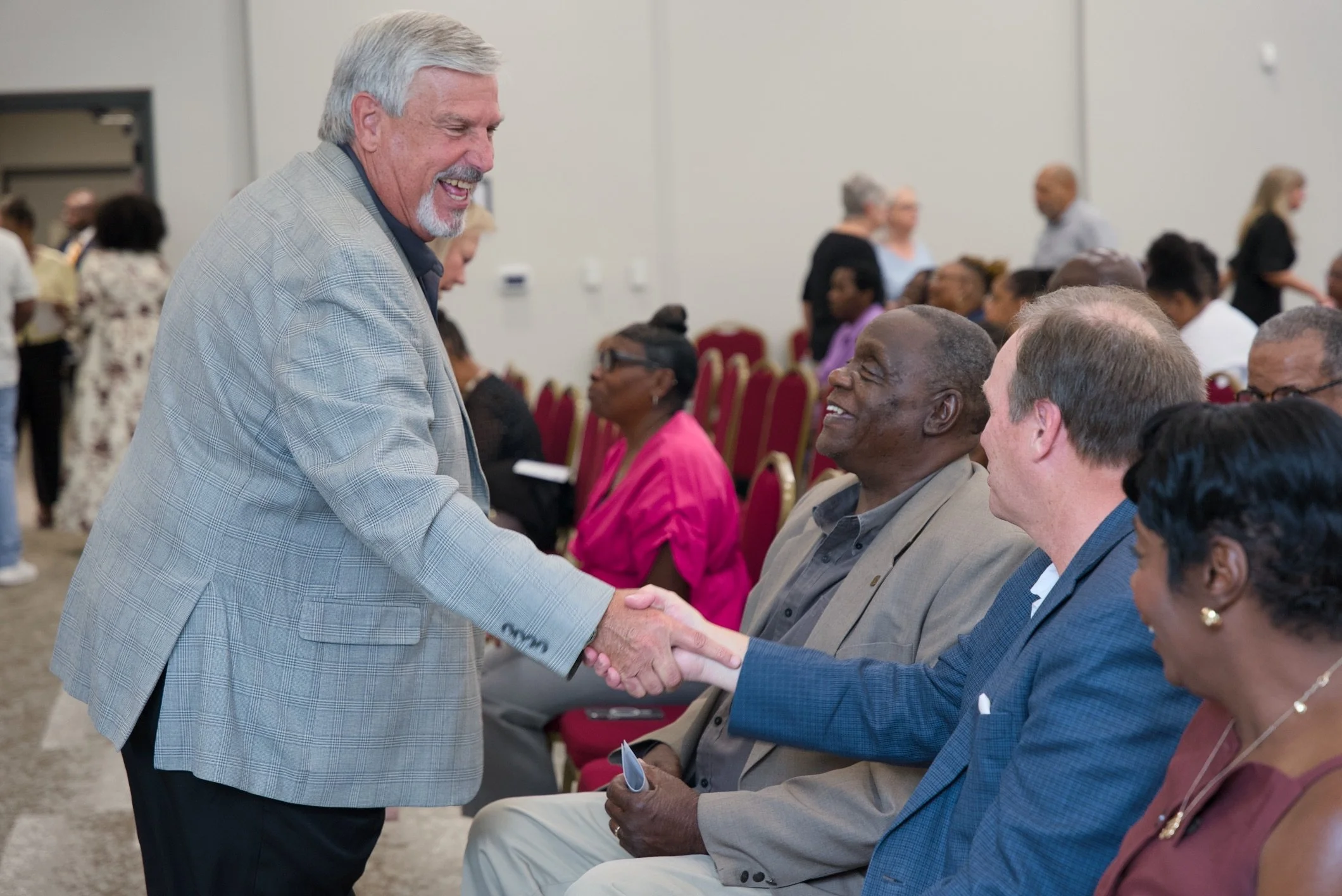 A man with gray hair and a beard in a gray plaid blazer shakes hands with a seated man during a formal event in a large room. Several people are seated and engaged in conversation in the background.