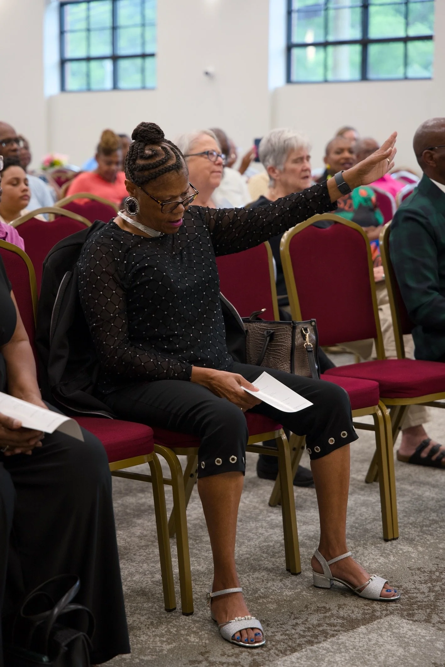 A group of women attending a conference or event, seated on red chairs with gold frames in a bright room with large windows. One woman in the foreground with braided hair and glasses has her eyes closed and hand raised.
