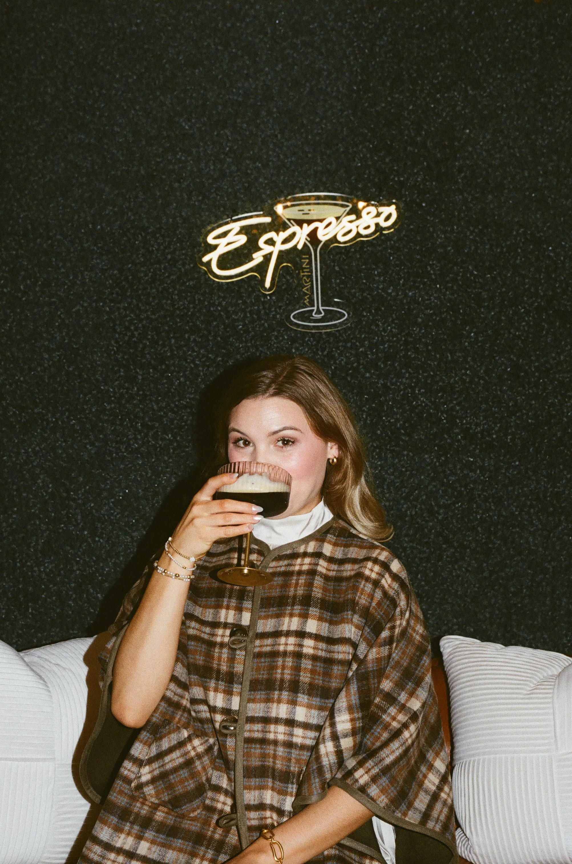 Young woman with light brown hair, wearing a plaid coat, sitting on a white sofa, drinking a dark cocktail, with a black textured wall and a neon sign reading 'Espresso' in the background.