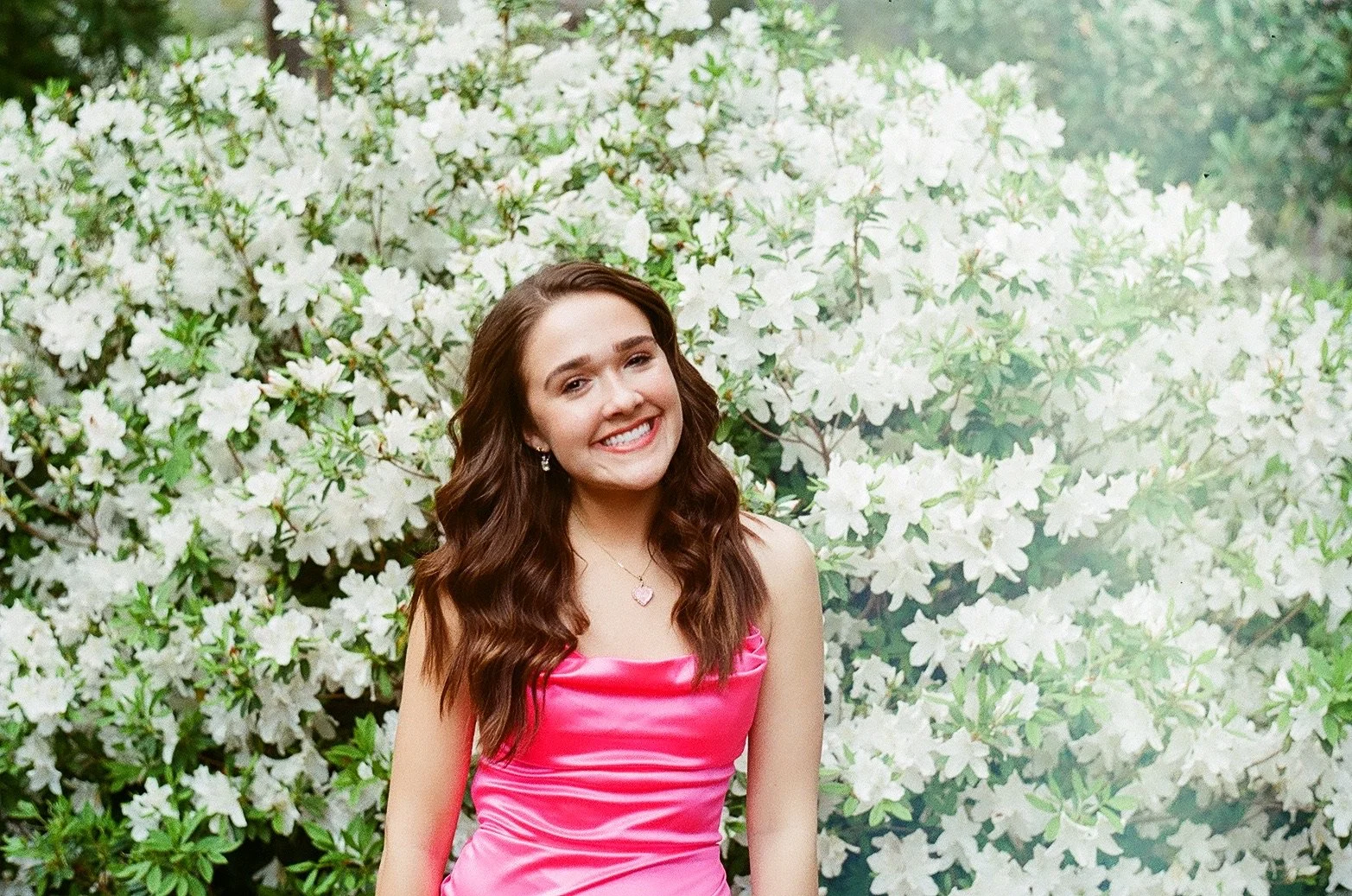 A young woman with long wavy brown hair, wearing a pink satin dress, jewelry, smiling, standing in front of a bush with white flowers.