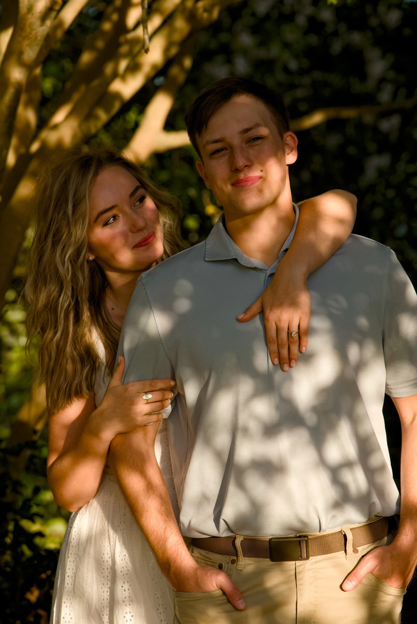 A young couple standing outdoors in a sunny setting, with the woman smiling and looking at the man, who is smiling at the camera. The woman's arm is wrapped around the man's shoulder, and they appear happy.