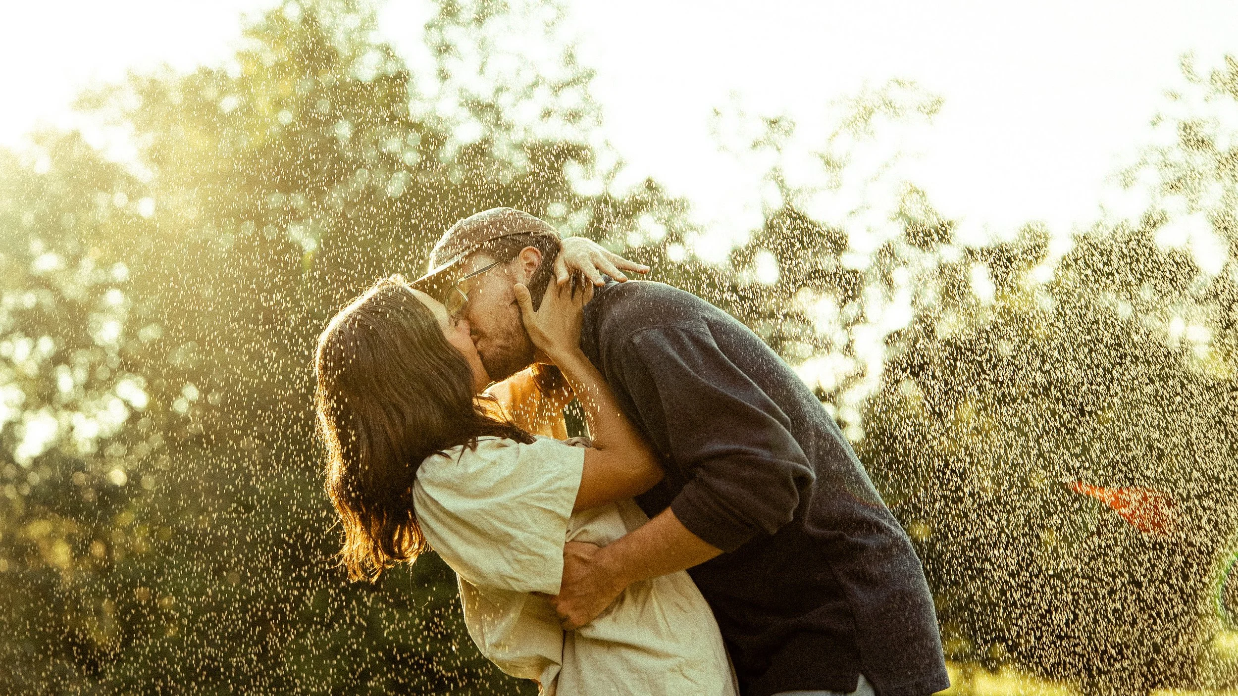 A couple kisses outdoors in warm sunlight with water droplets around them.