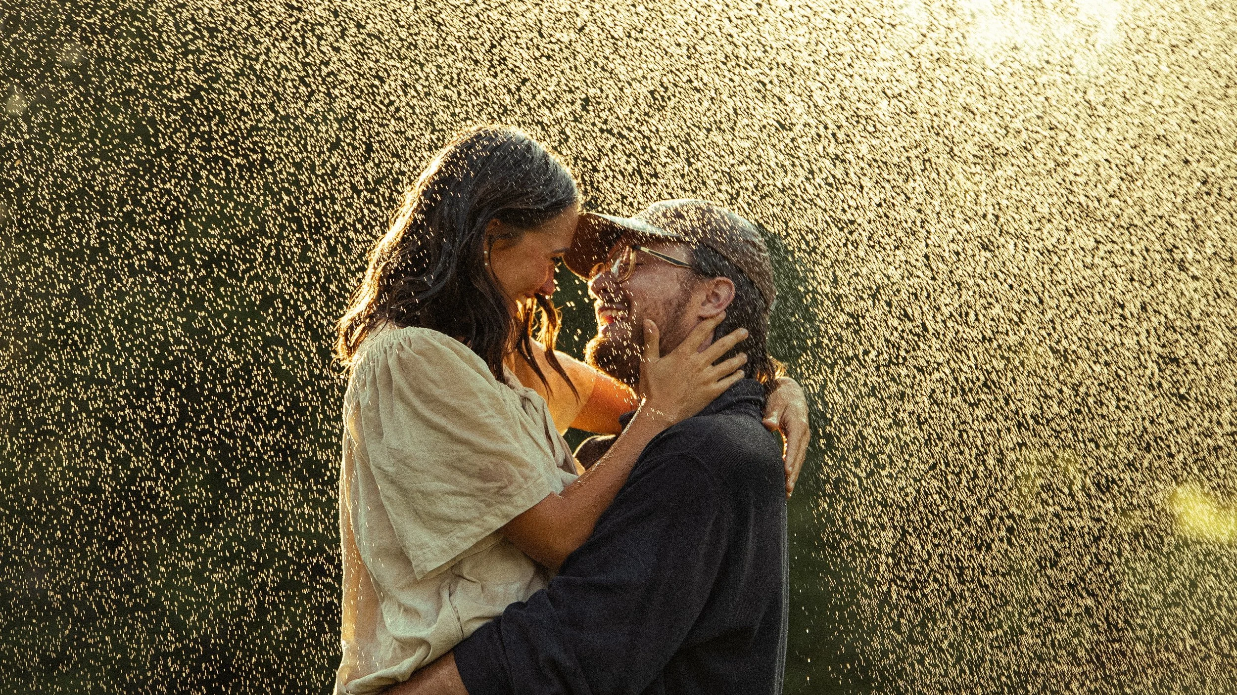 A couple embracing and smiling at each other outdoors with sunlight and particles surrounding them.