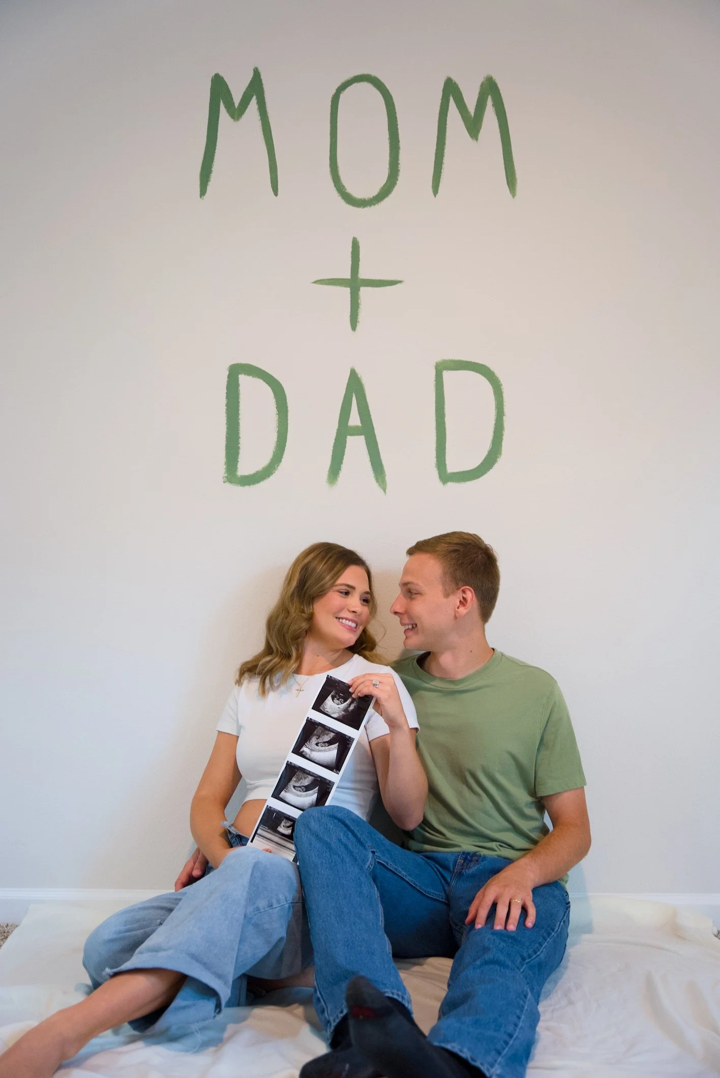 A couple sitting on a bed holding ultrasound photos, with a large sign on the wall behind them that reads 'MOM + DAD'