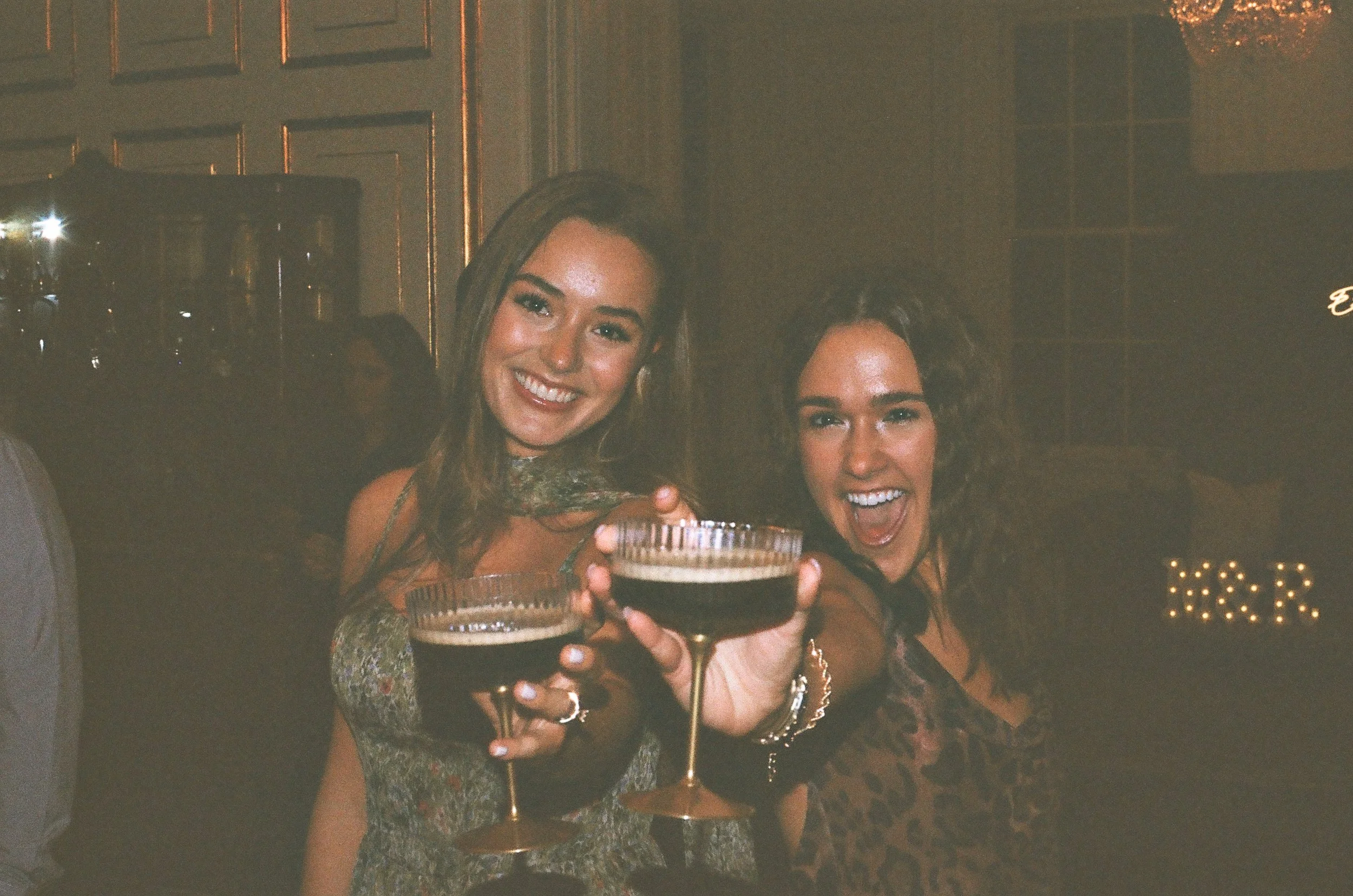 Two women smiling and holding dark cocktails in stemmed glasses, with a dimly lit, elegant background.