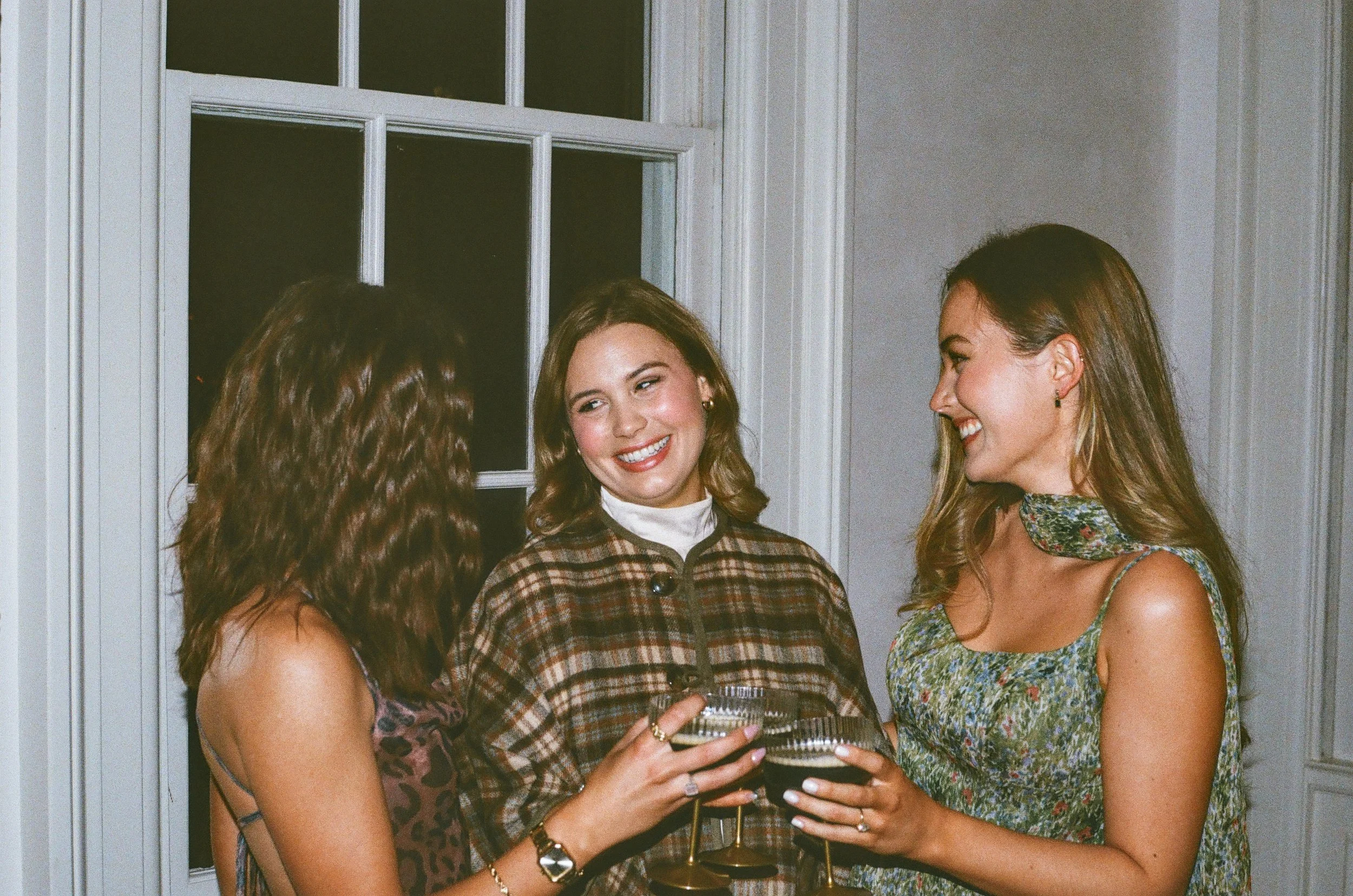 Three women enjoying drinks and conversation at a social gathering indoors near a window.