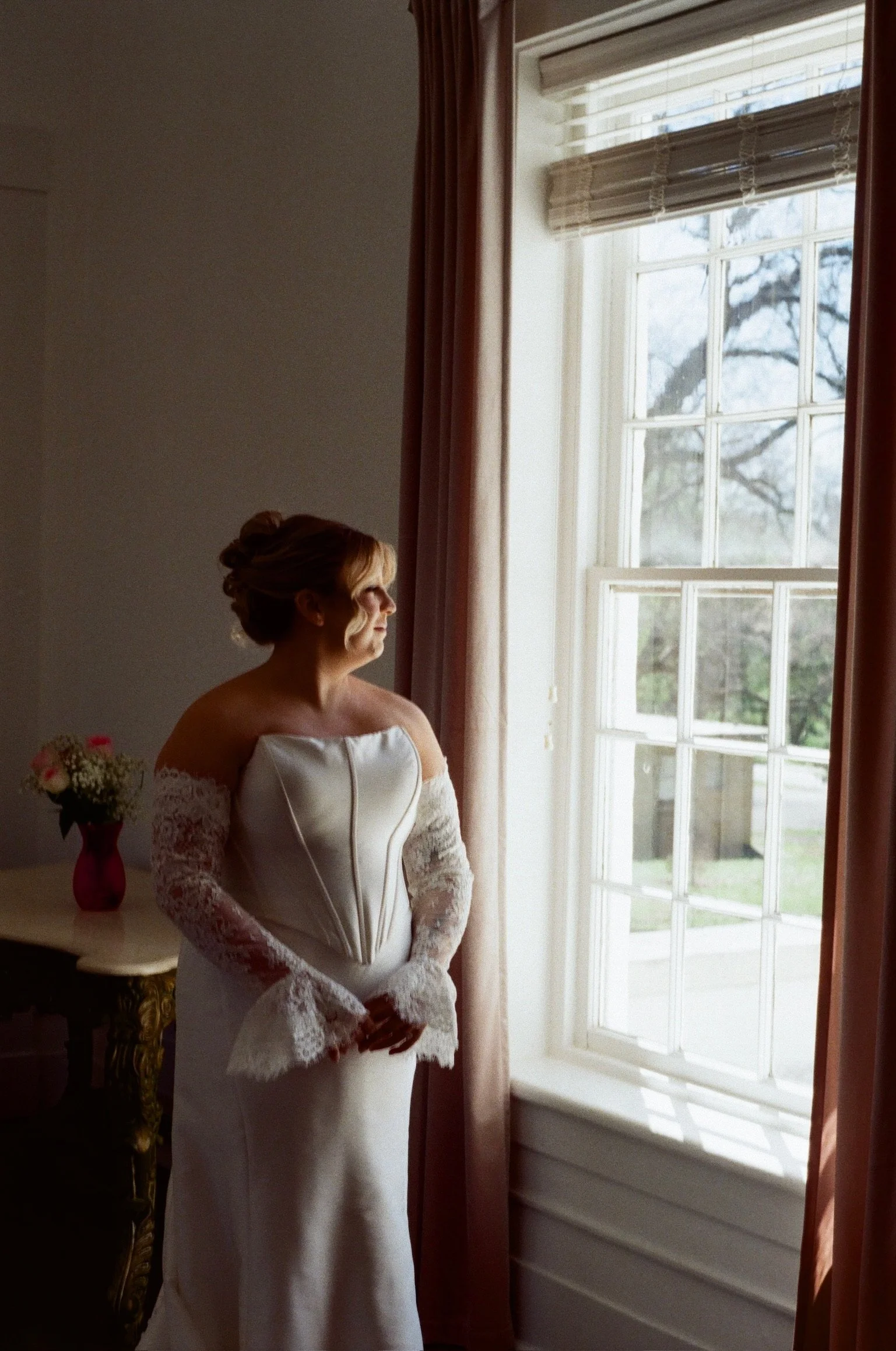 A woman in a wedding dress stands near a window with curtains, looking out.