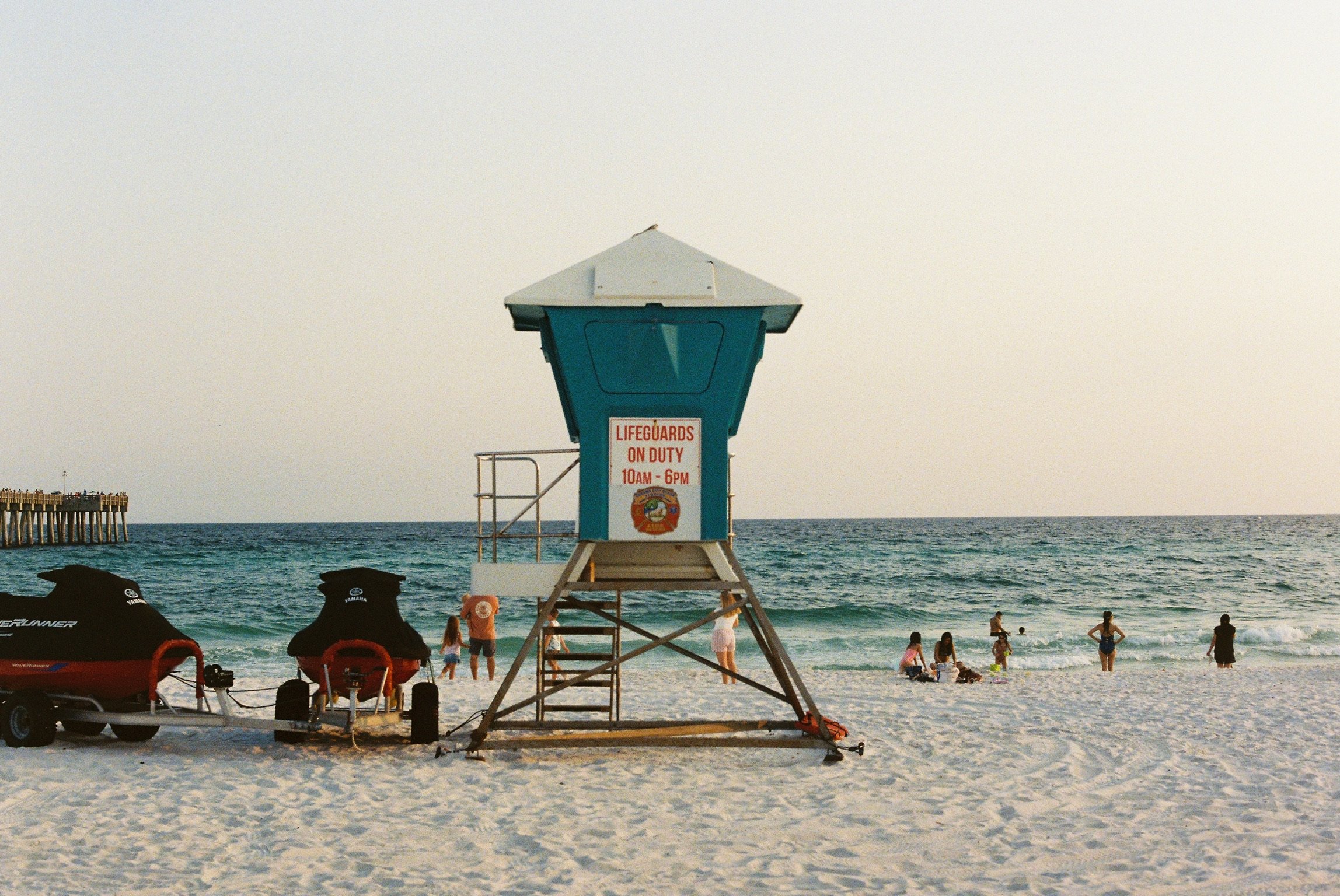 Lifeguard tower by the beach with a sign indicating lifeguards on duty from 10am to 6pm. People are swimming and relaxing in the water and on the sand, with a pier in the distance under a clear sky.