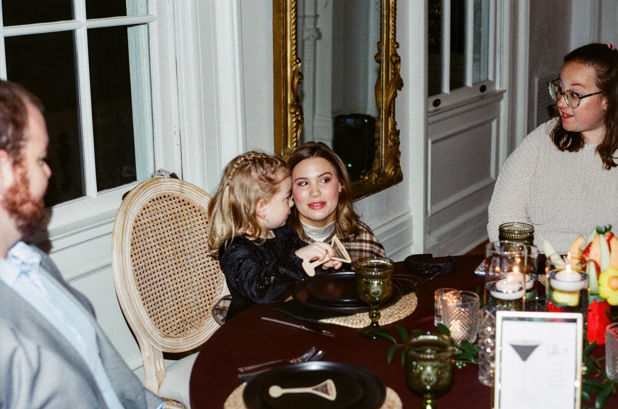 A group of women and girls sitting at a dinner table, engaging in conversation, with Christmas-themed decorations and food on the table, in a room with ornate gold-framed mirrors and white-paneled walls.