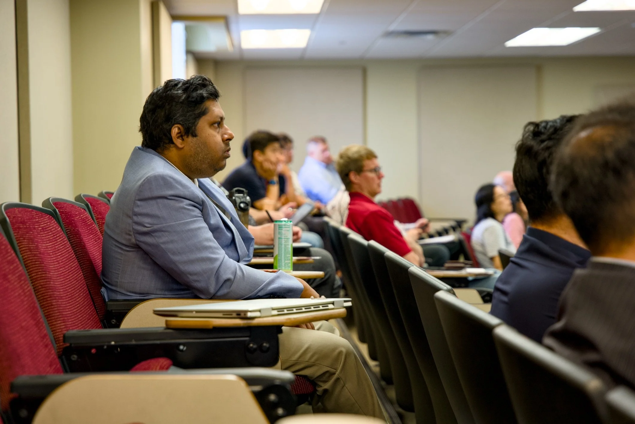 People attending a conference or seminar in a room with red chairs, some taking notes, focused on a speaker or presentation.