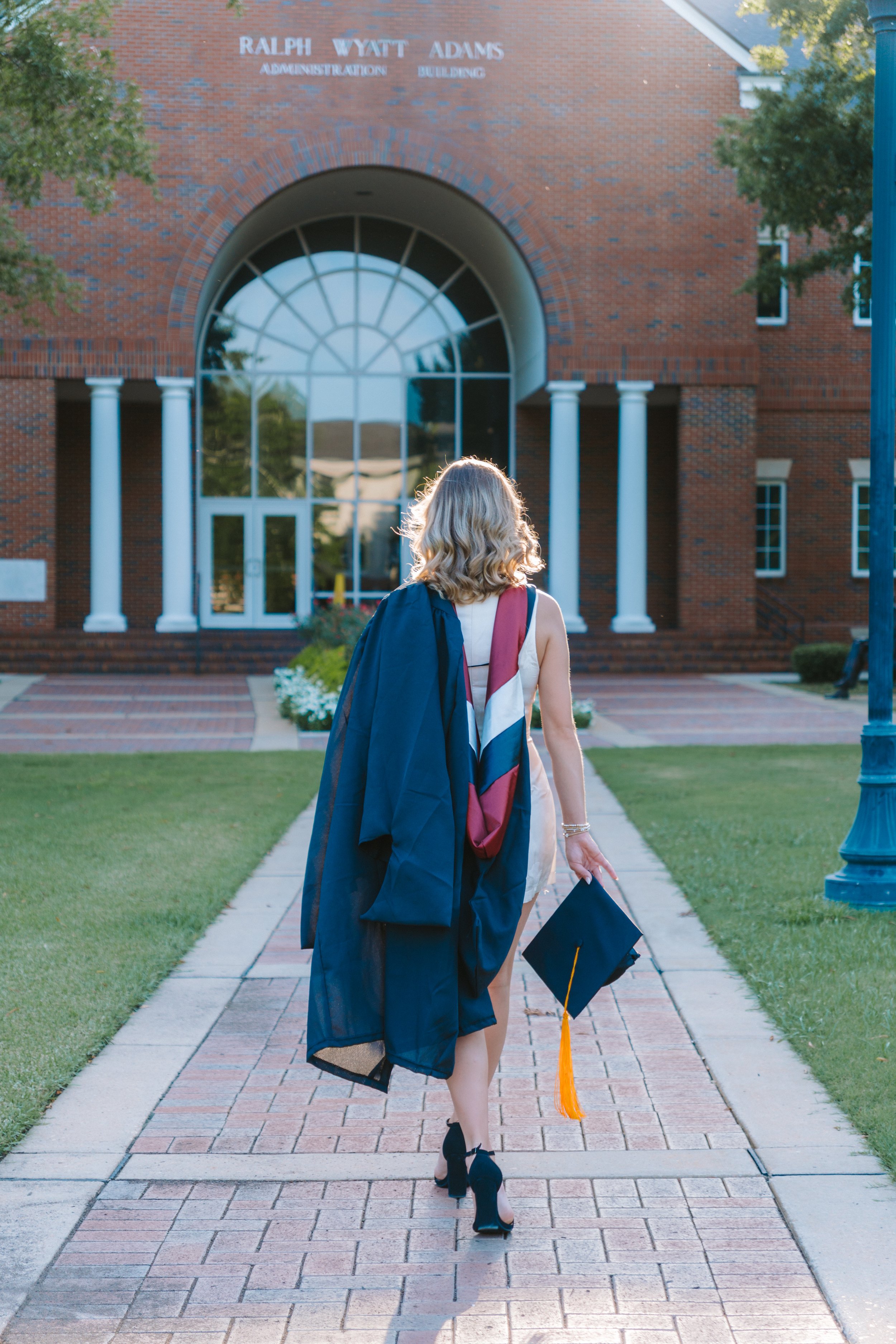 A woman wearing a graduation cap and gown walking towards a brick university building with an arched window and white columns.
