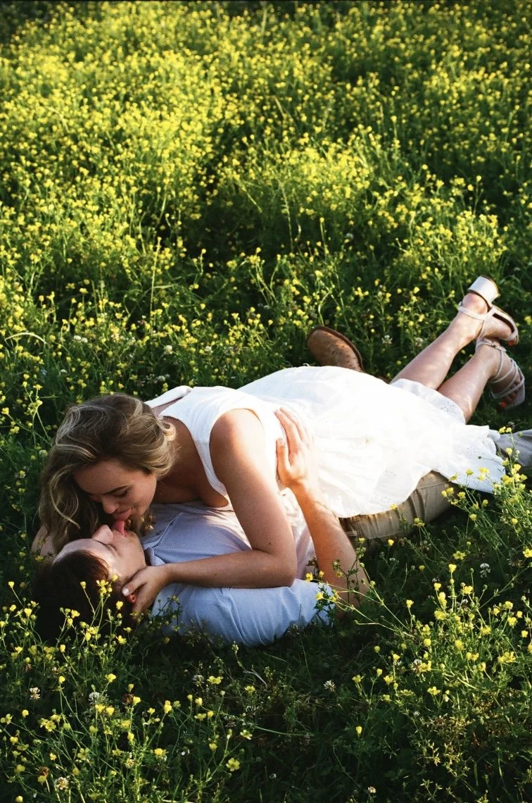 A couple lying in a field of yellow flowers, with the woman leaning over and kissing the man's forehead.