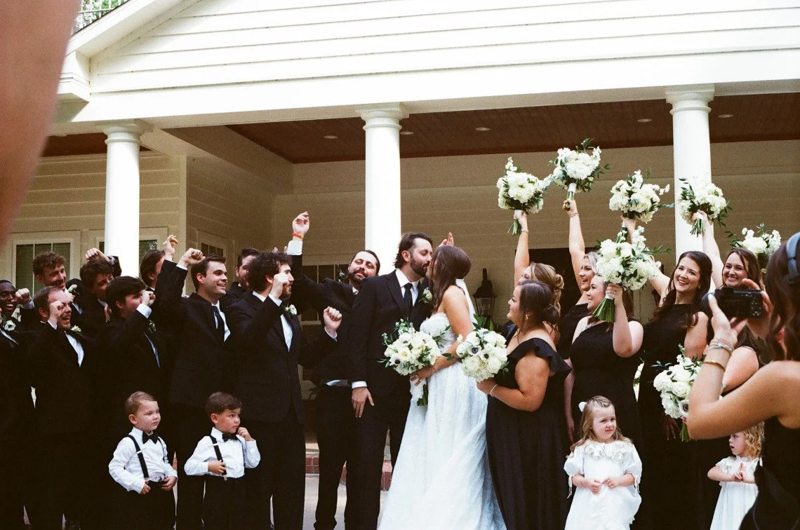 A wedding celebration with the bride and groom kissing, surrounded by friends and family, some holding bouquets, in front of a house with columns.